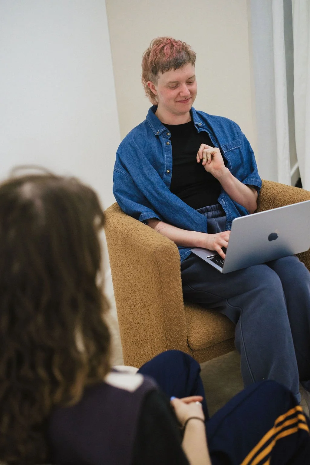 A person with short, pink hair and a blue denim shirt sitting in a brown armchair with a laptop on their lap, engaging in conversation with another person whose face is not visible in the foreground.