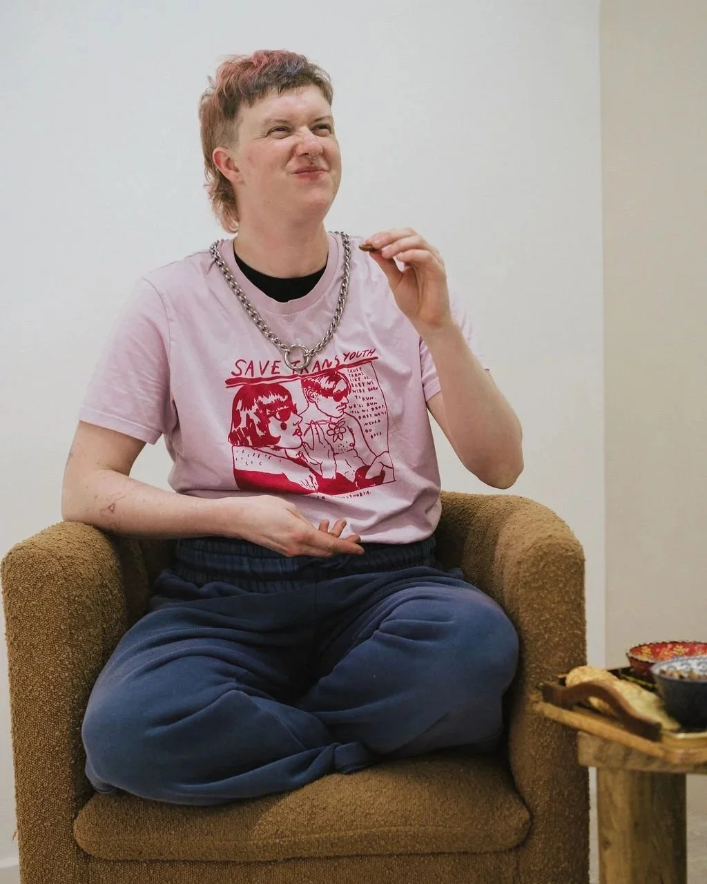 Person with short hair, wearing a pink graphic T-shirt with red line drawing and text, and dark blue pants, sitting cross-legged in a brown chair, holding a snack near their mouth with a slight smile.