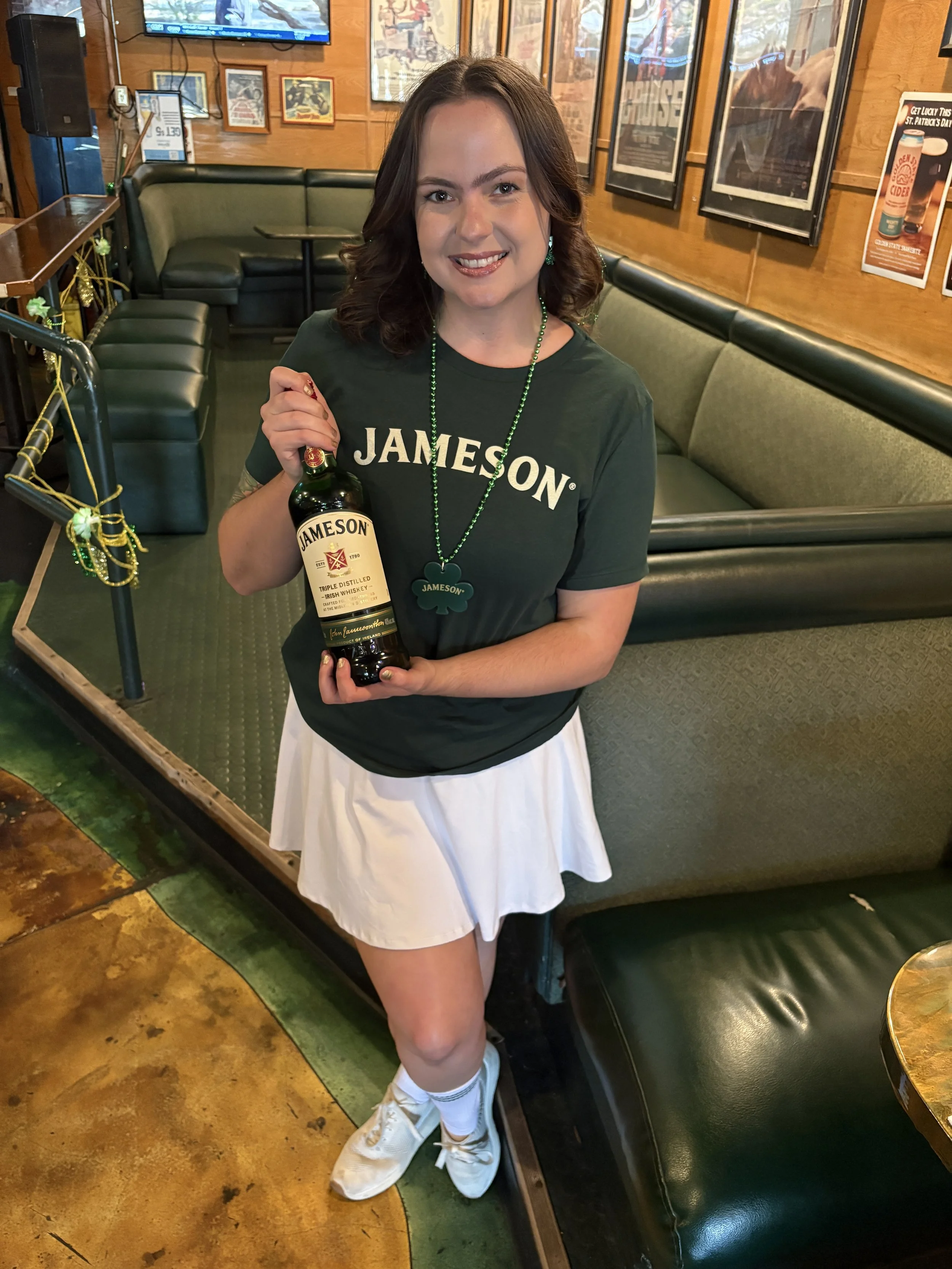 A young woman with shoulder-length brown hair smiling, wearing a Dark green Jameson T-shirt, a white skirt, holding a bottle of Jameson Irish whiskey inside a bar or restaurant. Working as a brand ambassador for an activation. Promotional modeling.