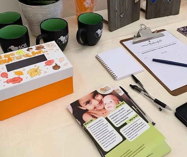 Desk with three black and green mugs, a tissue box, a small notebook, pens, a clipboard with paper, and a brochure showing a woman and a baby.