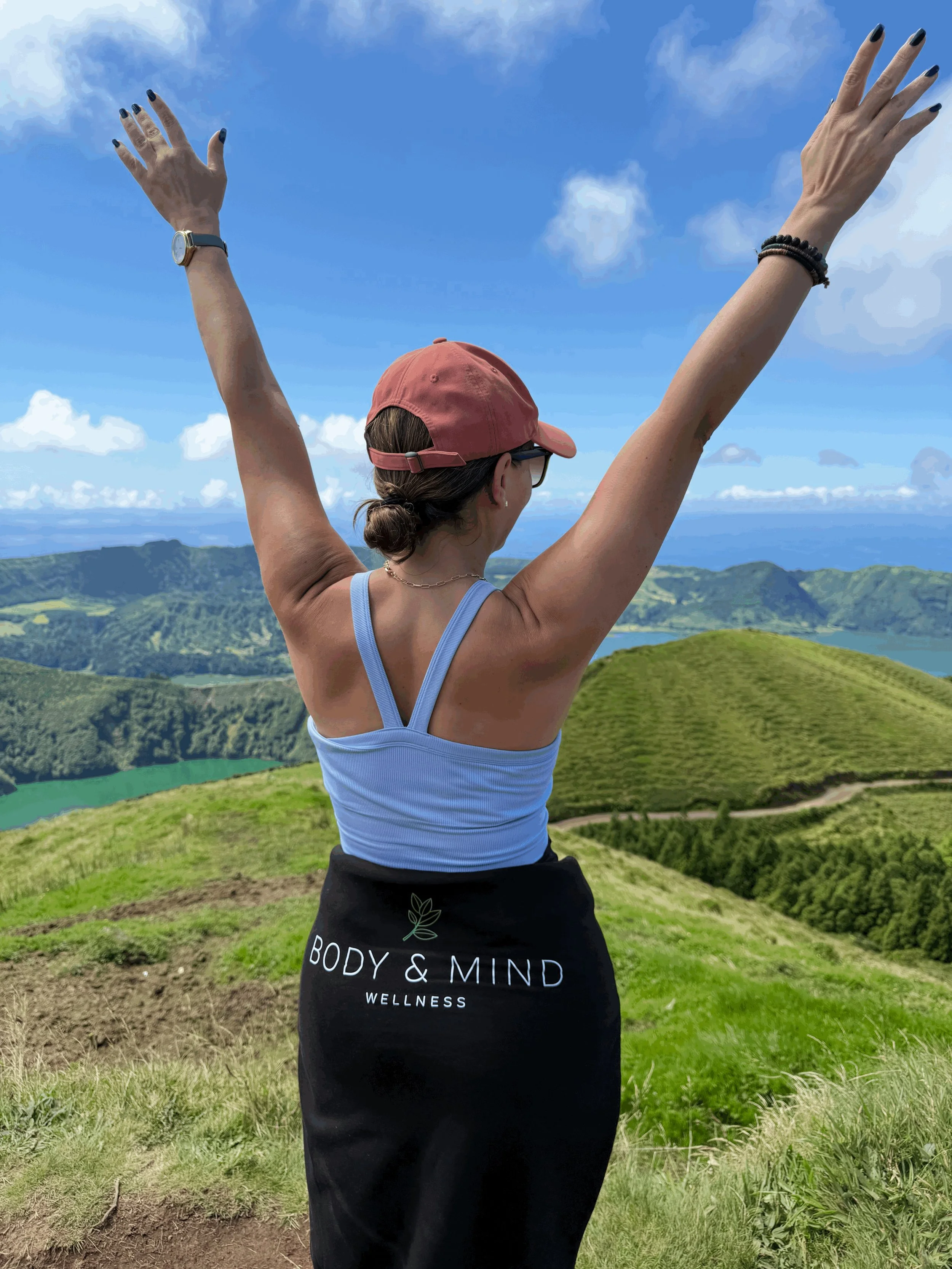 Woman standing on a grassy hill with arms raised in front of a scenic landscape of rolling green hills.