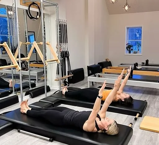 Two women doing Pilates exercises on reformer machines in a fitness studio with mats and exercise equipment.