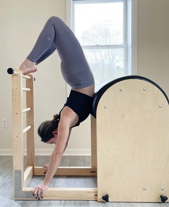 Person performing a backbend yoga pose on a wooden Pilates or exercise machine in a bright room with a window.