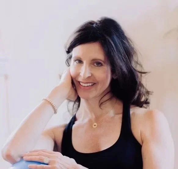 A woman with shoulder-length dark hair smiling, wearing a black tank top, and resting her head on her left hand.