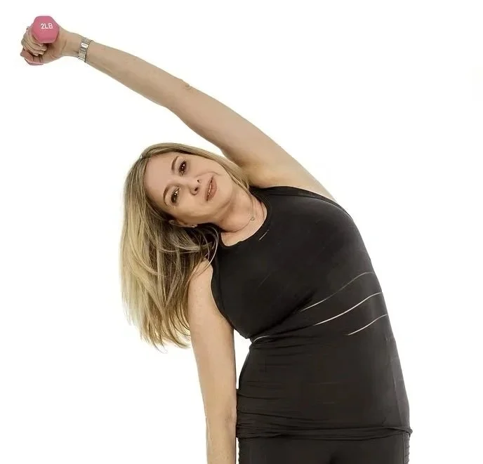 A woman in athletic clothing stretching with a pink 2-pound dumbbell in her right hand, leaning sideways with her head tilted and smiling.