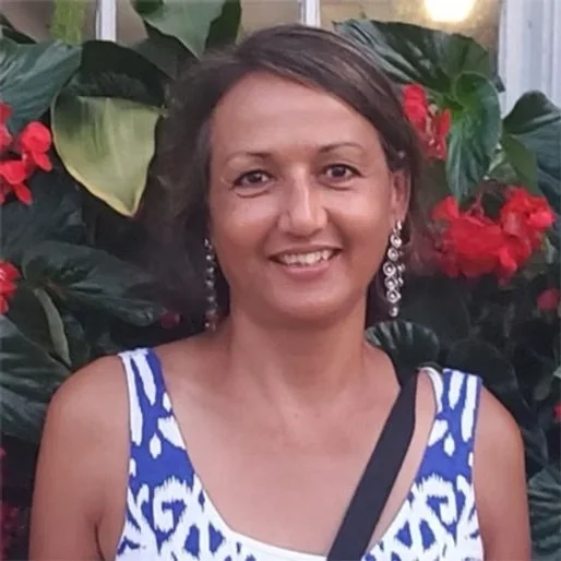 A woman with short dark hair smiling, wearing dangle earrings and a blue and white patterned sleeveless top, standing in front of green leaves and red flowers.