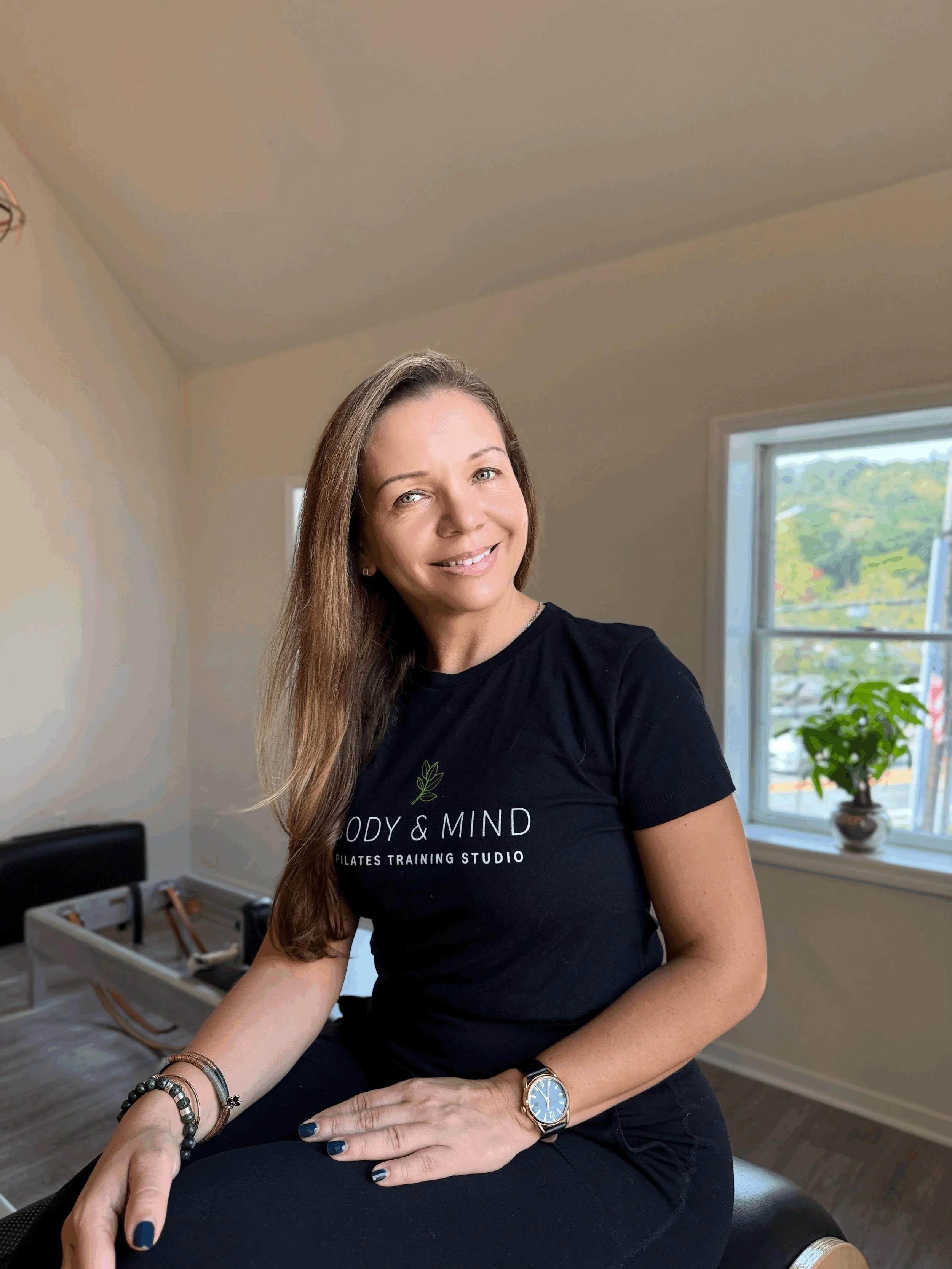 A woman with long brown hair and light skin smiling and sitting on a chair in a room with natural light coming through the window. She is wearing a black T-shirt with a logo reading 'Body & Mind Pilates Training Studio,' a watch, and bracelets. There is a potted plant on the windowsill and an open suitcase in the background.