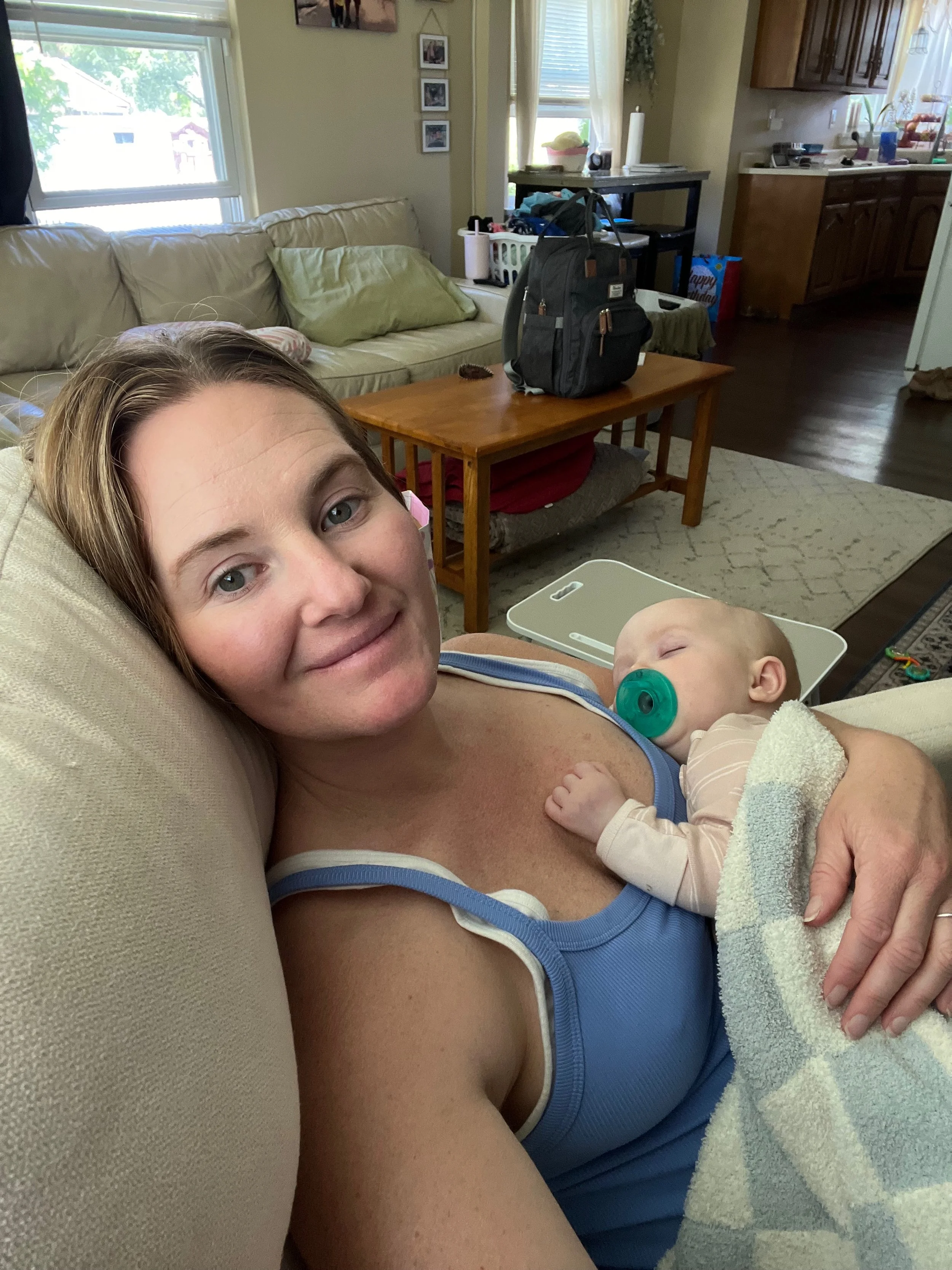 A woman is lying on a beige couch holding a sleeping baby with a pacifier in its mouth. The woman is smiling at the camera, wearing a blue tank top. The background shows a living room with a window, a coffee table, and a kitchen area.