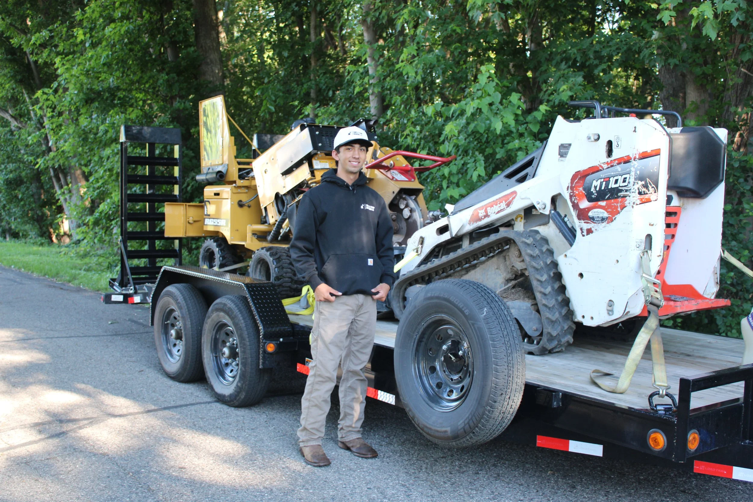 A man standing in front of a flatbed trailer carrying construction equipment, including a yellow Bobcat compact loader and a white mini excavator, with woods in the background.