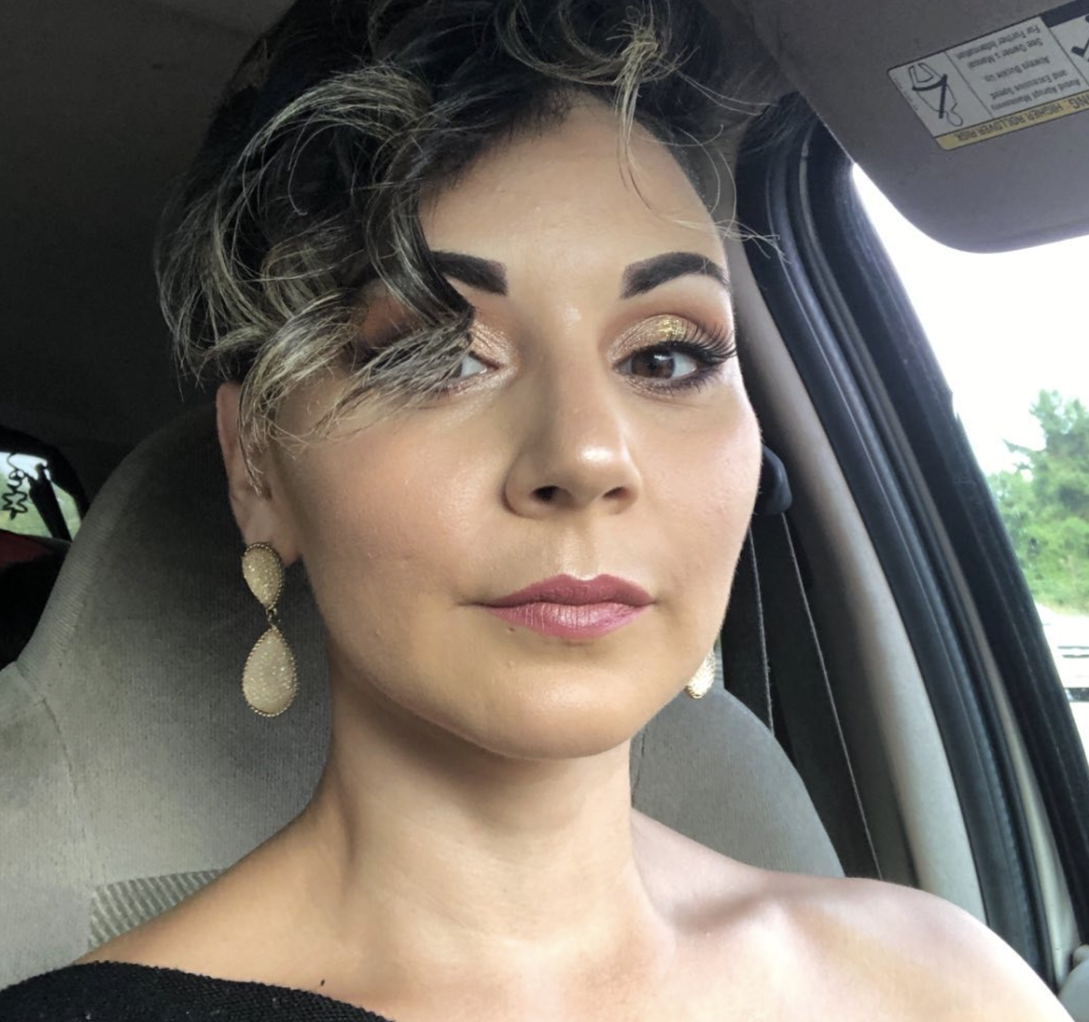 A woman with short, wavy brunette hair, wearing gold earrings and makeup, sitting inside a car with a view of trees outside.
