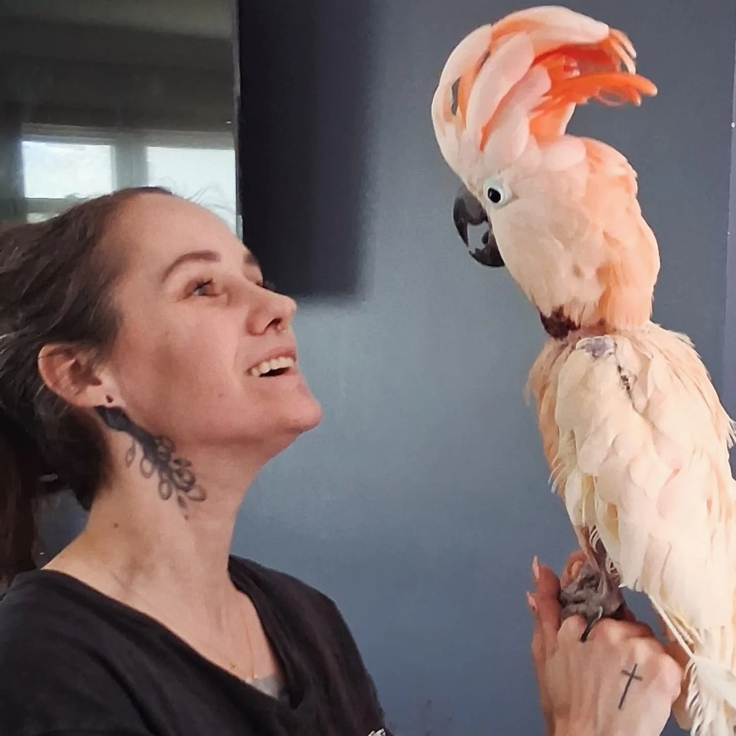 A woman with a tattoo on her neck is smiling and looking at a pink cockatoo perched on her hand.
