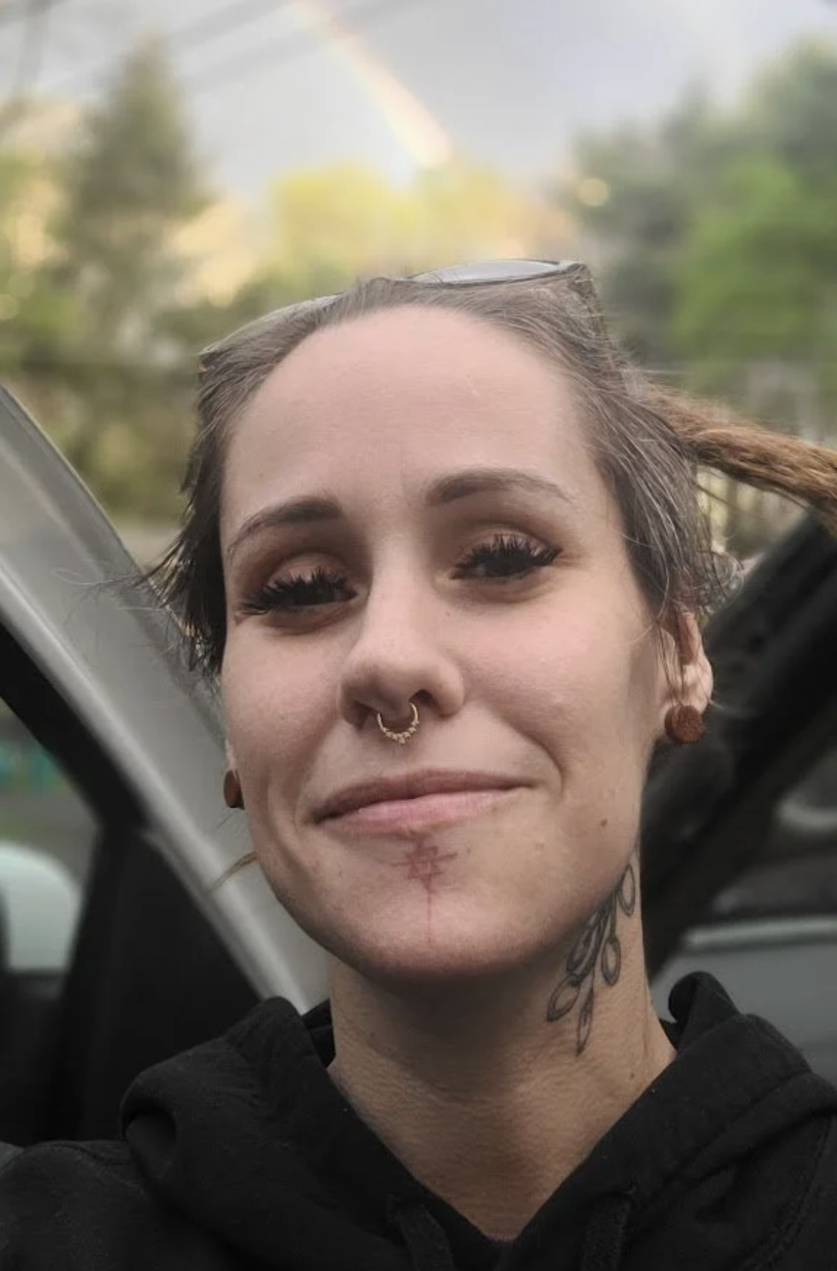 A woman with brown hair, eye makeup, a septum piercing, a tattoo on her neck, and earrings, smiling with a rainbow in the background.