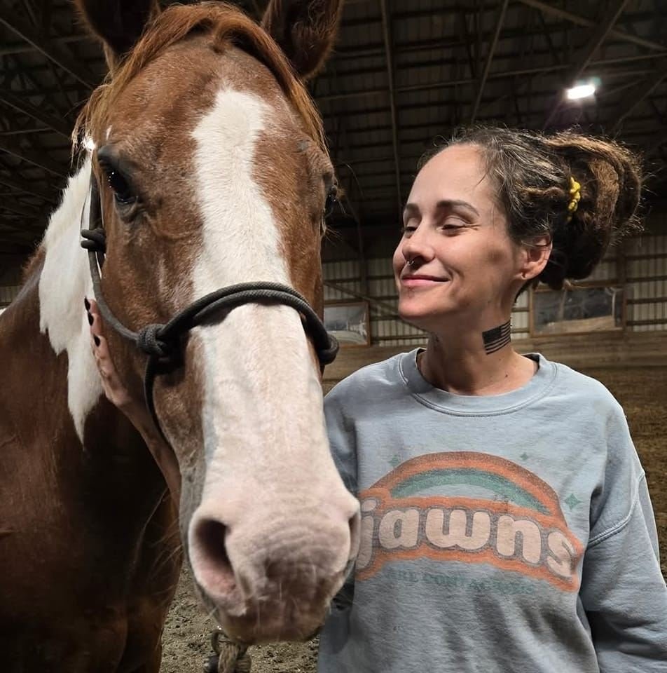 A woman smiling happily next to a large brown and white horse inside a barn.