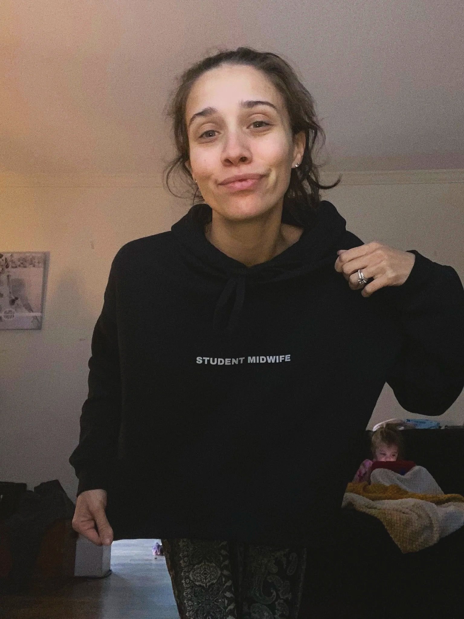Young woman wearing a black hoodie with 'Student Midwife' written on it, standing indoors.
