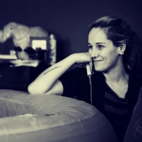 A woman with long hair, sitting indoors, resting her chin on her hand, smiling softly.