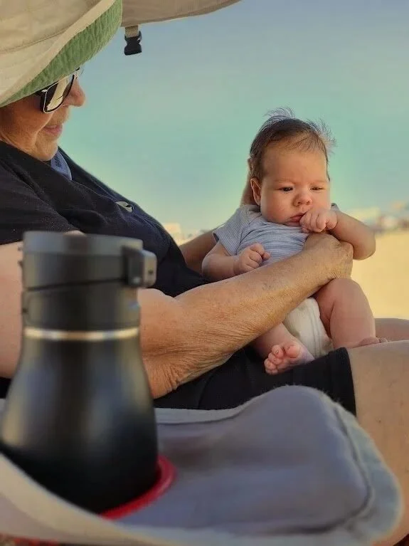 An older person holding a baby on a beach under a canopy, with a black water bottle in the foreground.