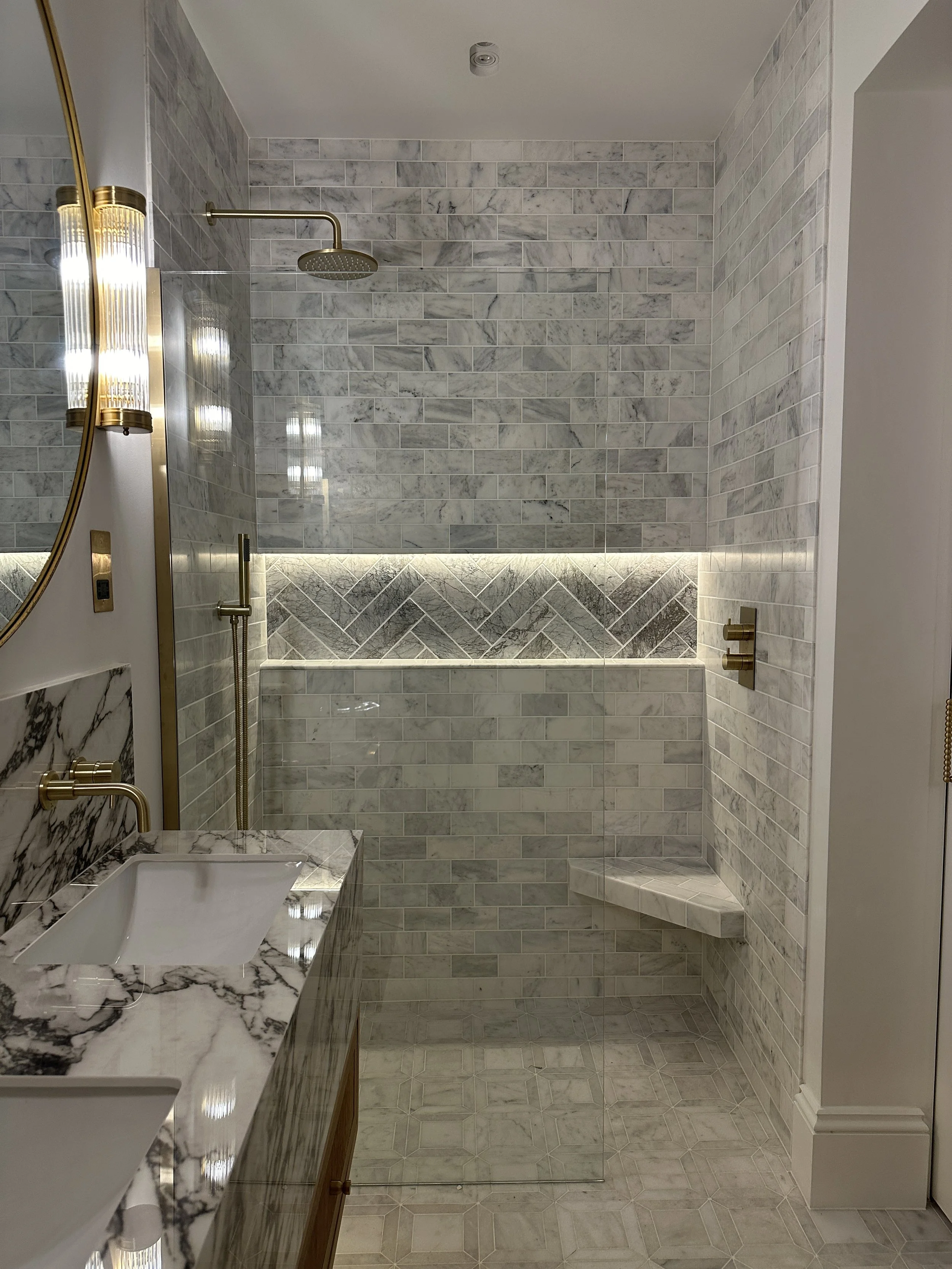 Modern bathroom with a marble countertop and dual sinks, a large mirror, gold fixtures, a walk-in shower with marble tiles, a built-in bench, and intimate LED lighting.