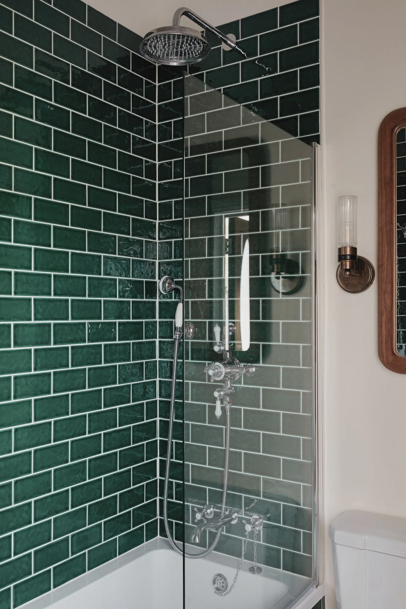 A shower area with dark green subway tiles, a rain shower head, a handheld shower, and a glass shower divider. Part of a mirror, wall sconce, and toilet are visible outside the shower.