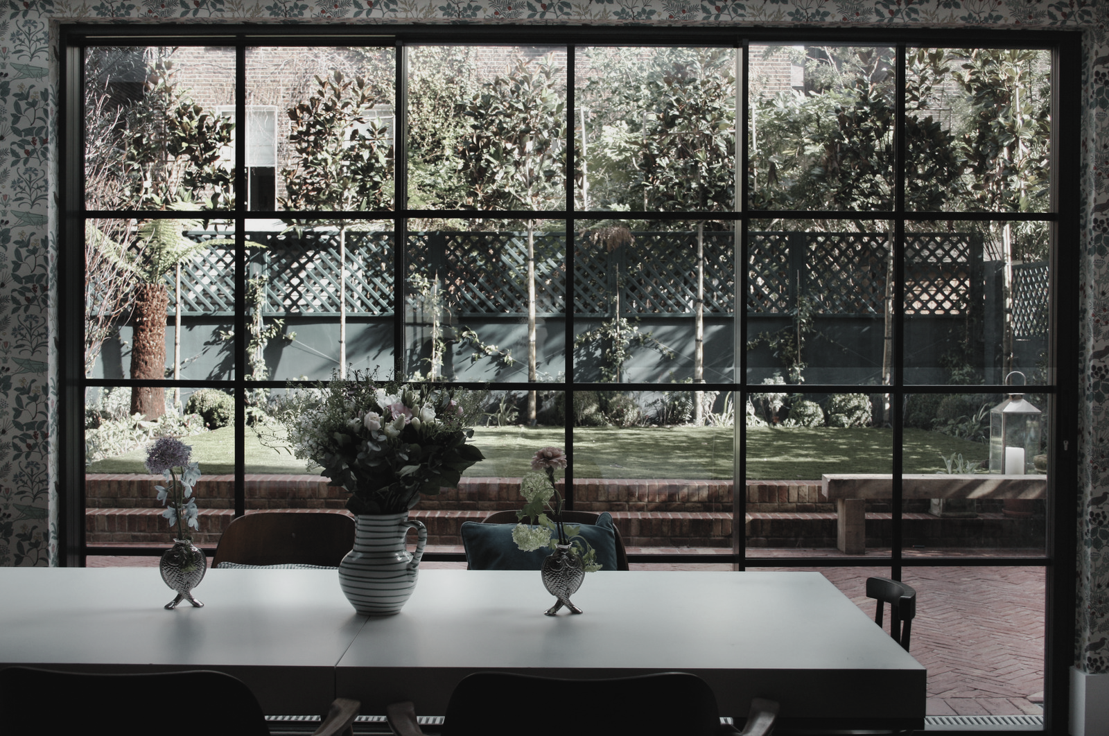 Dining room table with vases and a flower arrangement, overlooking a garden through large glass windows.