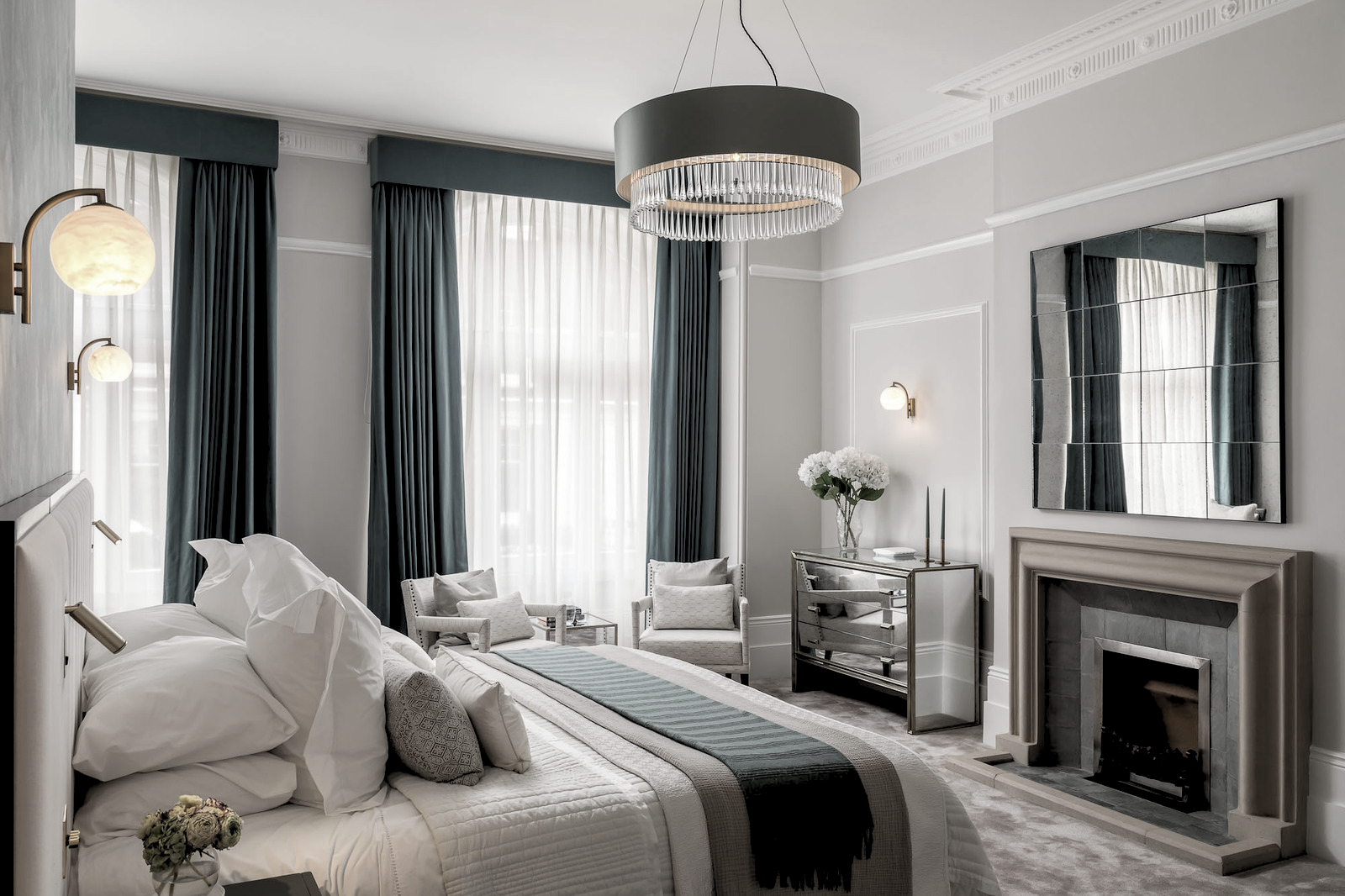 Elegant bedroom with white walls, blue curtains, a large bed with white bedding and grey pillows, a mirror above the fireplace, and a modern chandelier.