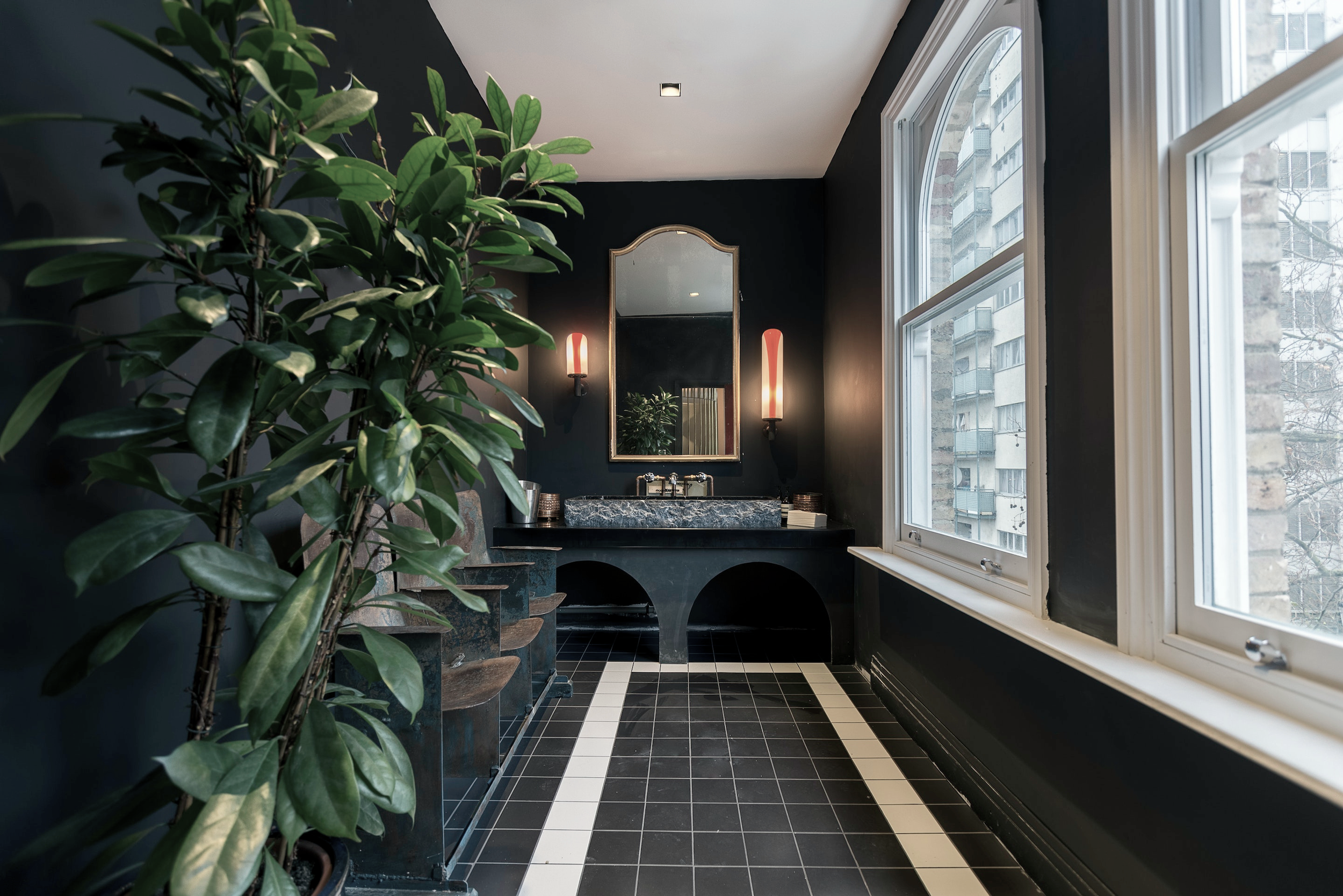 Modern bathroom with black walls, large windows, a black and white tiled floor, a vanity with a marble countertop, a mirror, and wall sconces.
