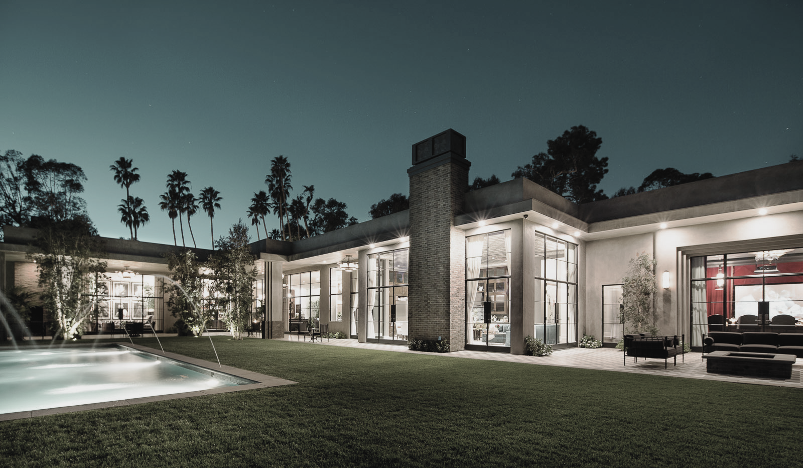 Night view of a modern house with large glass windows and a lit backyard, including a fountain and outdoor seating area, with tall palm trees in the background.