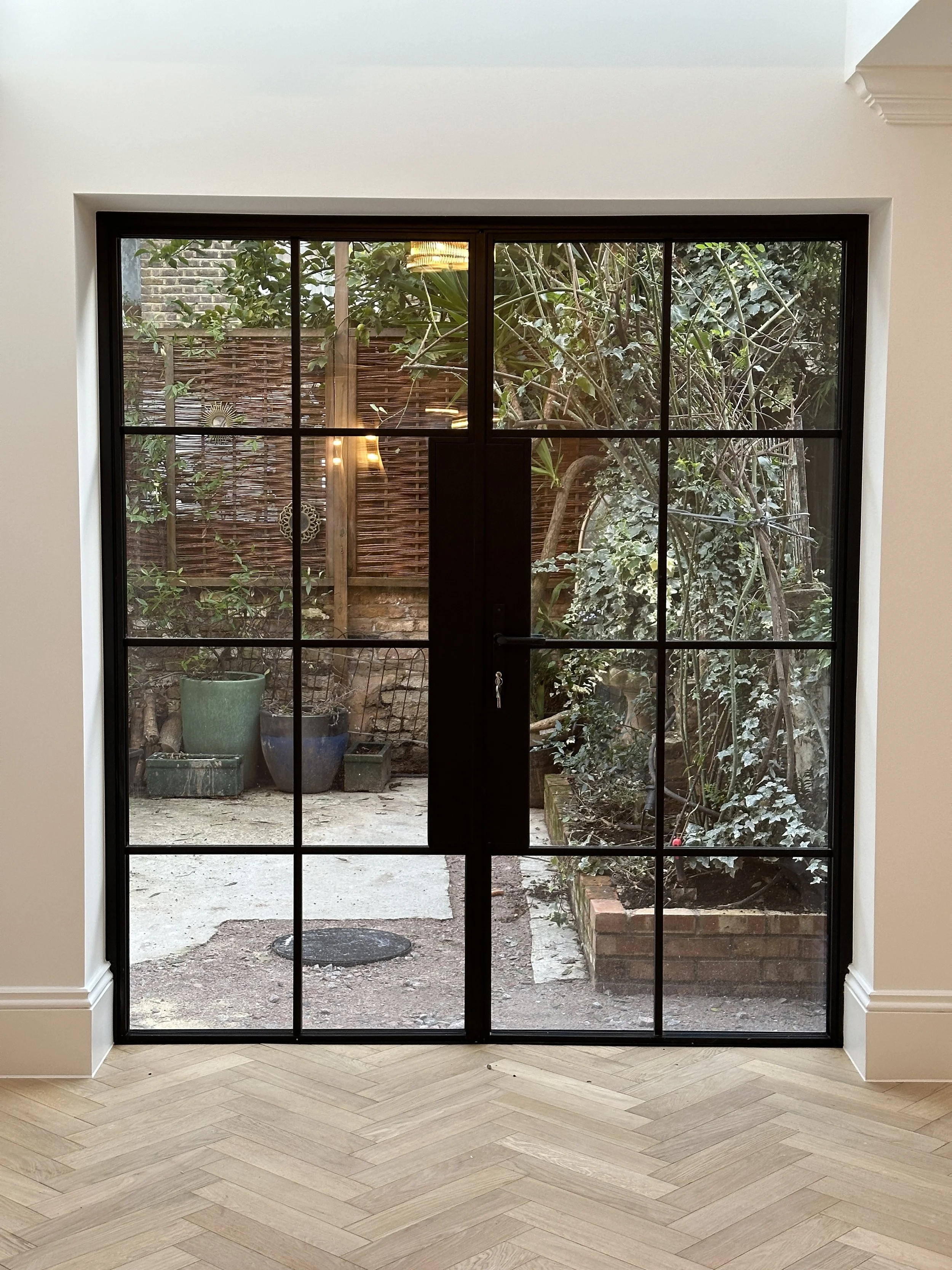 View through large black-framed glass door to small outdoor garden with potted plants, trees, and a brick-bordered flower bed.