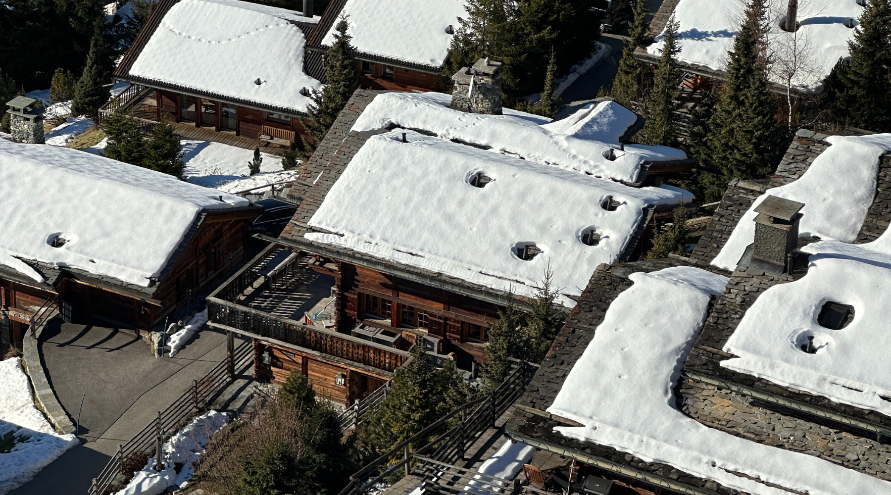 Aerial view of snow-covered rooftops of wooden houses in a mountain area, with trees and a road visible.