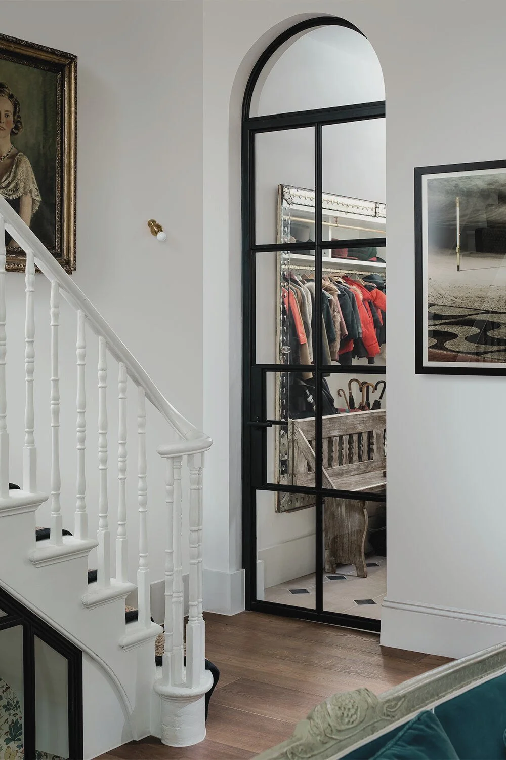 Interior view of a home showing a staircase with white railing, a framed portrait hanging on the wall, a large black-framed glass door leading to a walk-in closet with clothing and umbrellas, and a decorative framed artwork on the wall.