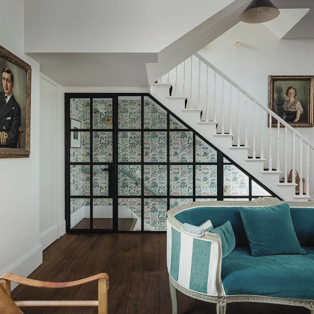 Interior of a living room featuring a staircase with white railings, floral wallpaper behind a black-framed glass door, a green velvet sofa with pillows, and framed vintage portraits on the wall.