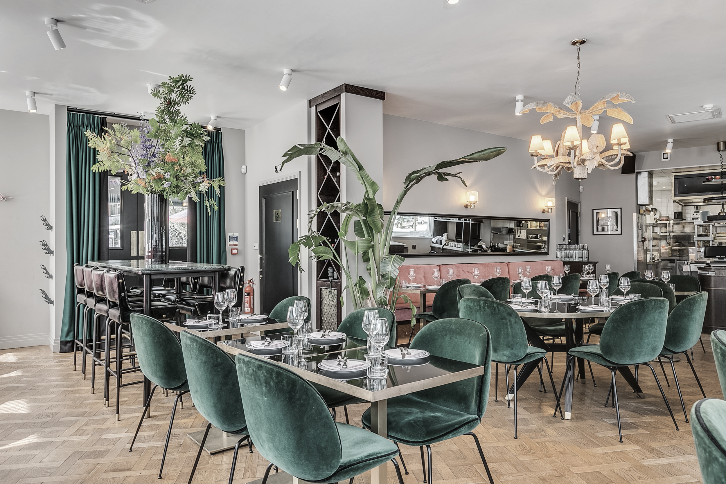 Elegant restaurant interior with green velvet chairs, tables set with glasses and plates, large floral arrangement, pink cushioned banquette, wood floor, chandelier, and open kitchen area in background.