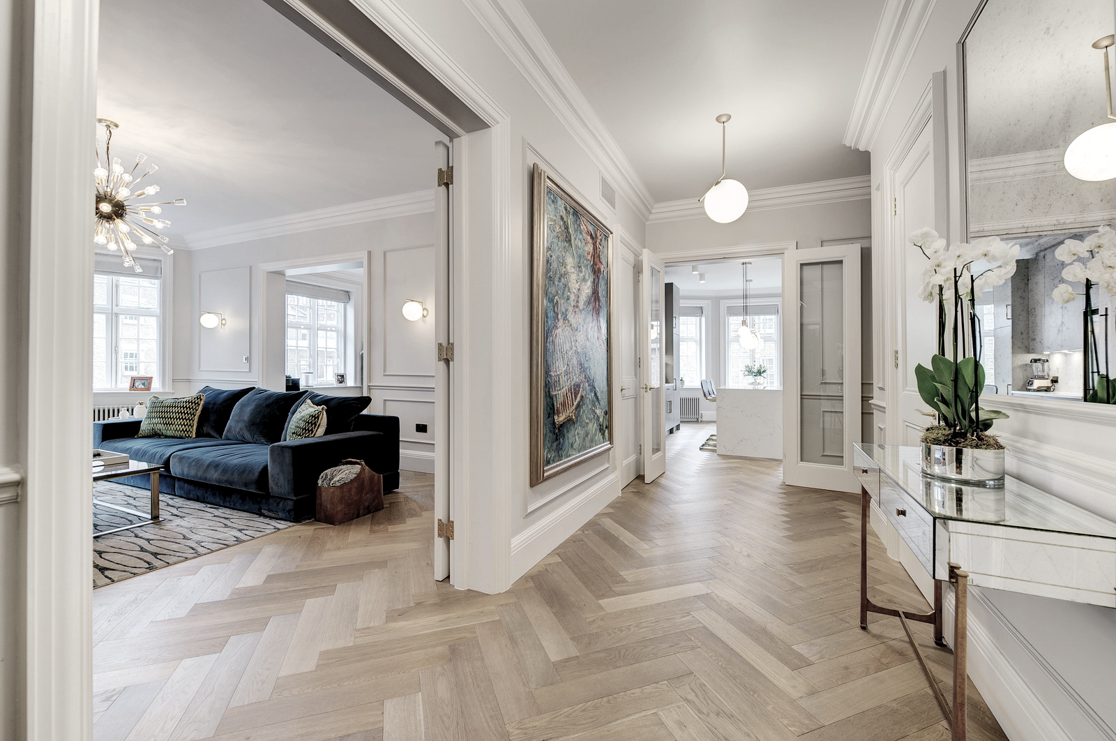 Bright modern apartment interior with a hallway leading to living room and kitchen, featuring white walls, wooden flooring, artwork, and light fixtures.