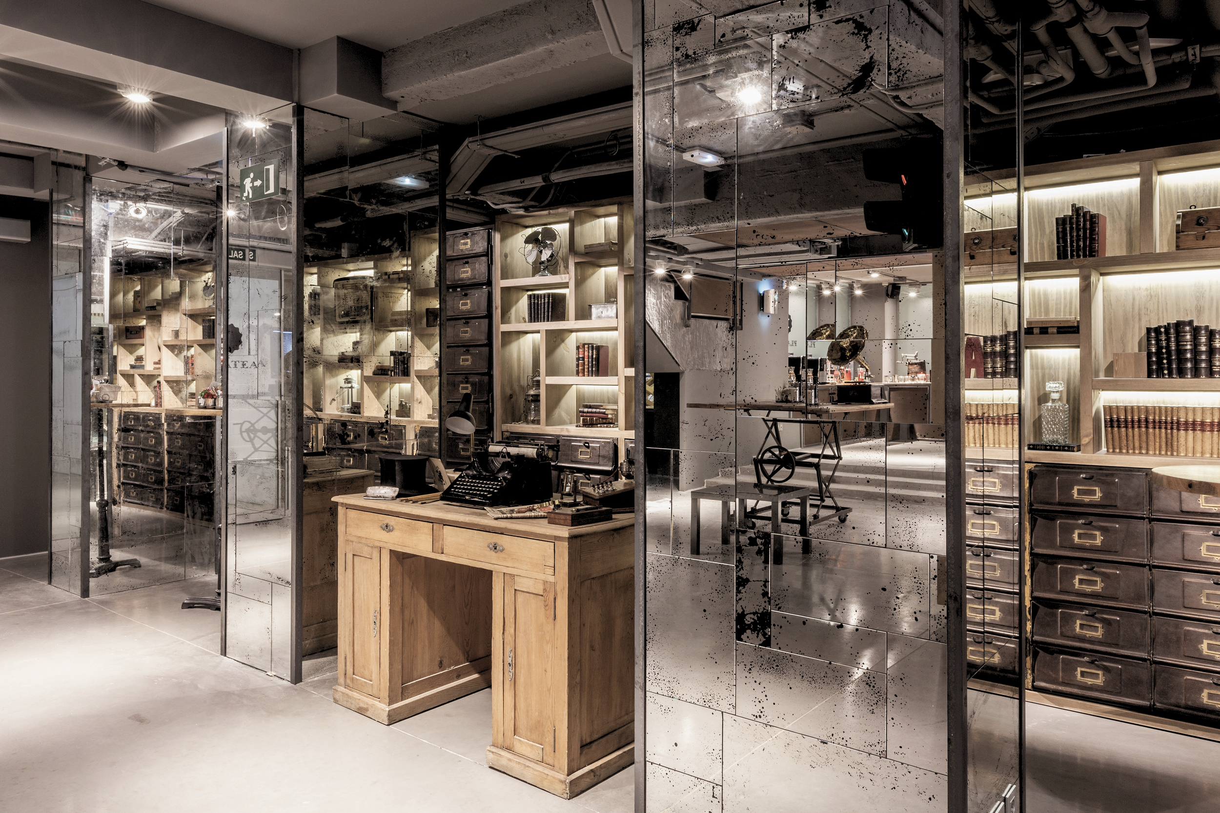 Interior of a trendy, industrial-style office or retail space with glass walls, wooden furniture, and metal shelves filled with books and decorative items.