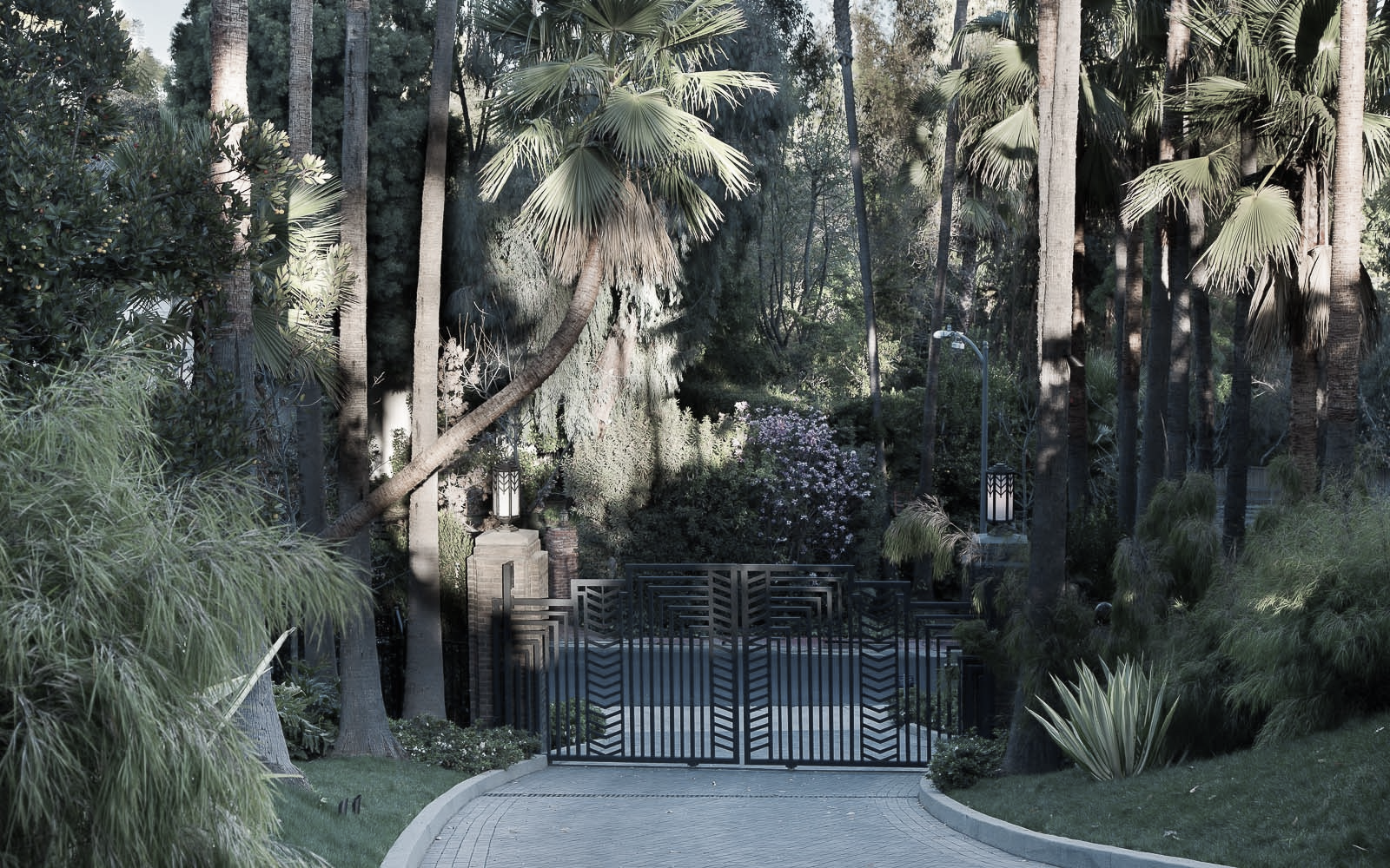 A gated entrance to a lush, tropical garden with tall trees, palm trees, and various shrubs and flowering plants, under dappled sunlight.