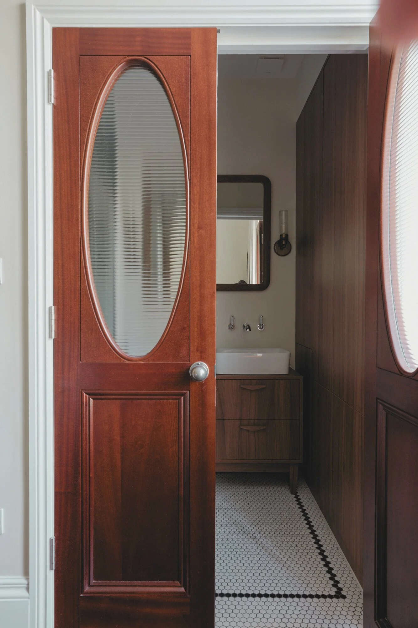 View of a bathroom with a wooden door partially open, revealing a sink, mirror, and wall-mounted faucet inside.