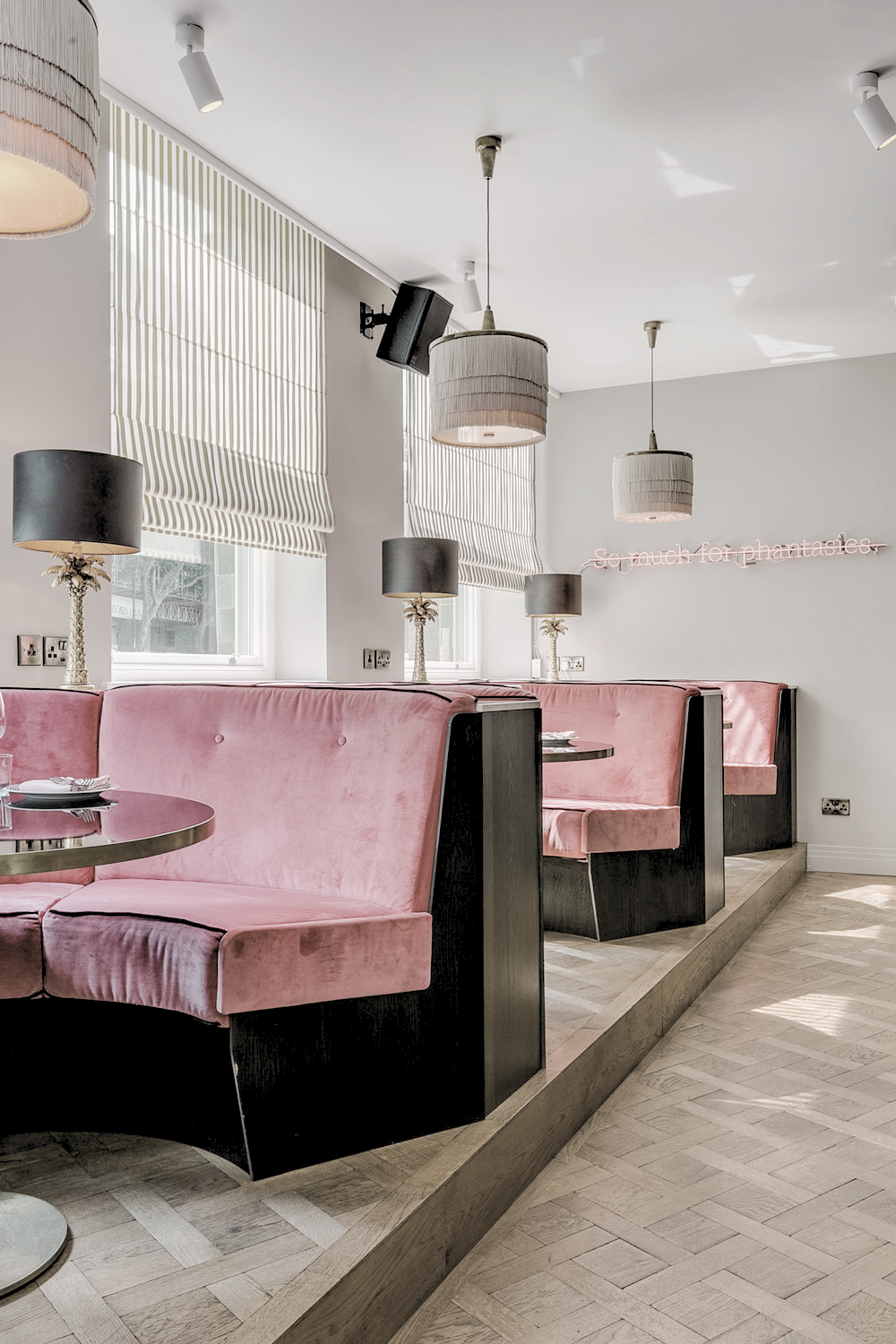 Interior of a modern cafe with pink velvet upholstered booths, black tables, black lamps with palm tree bases, and white ceiling lights. There are striped blinds on windows and a pink neon sign on the wall that says 'so much for phantasy'.