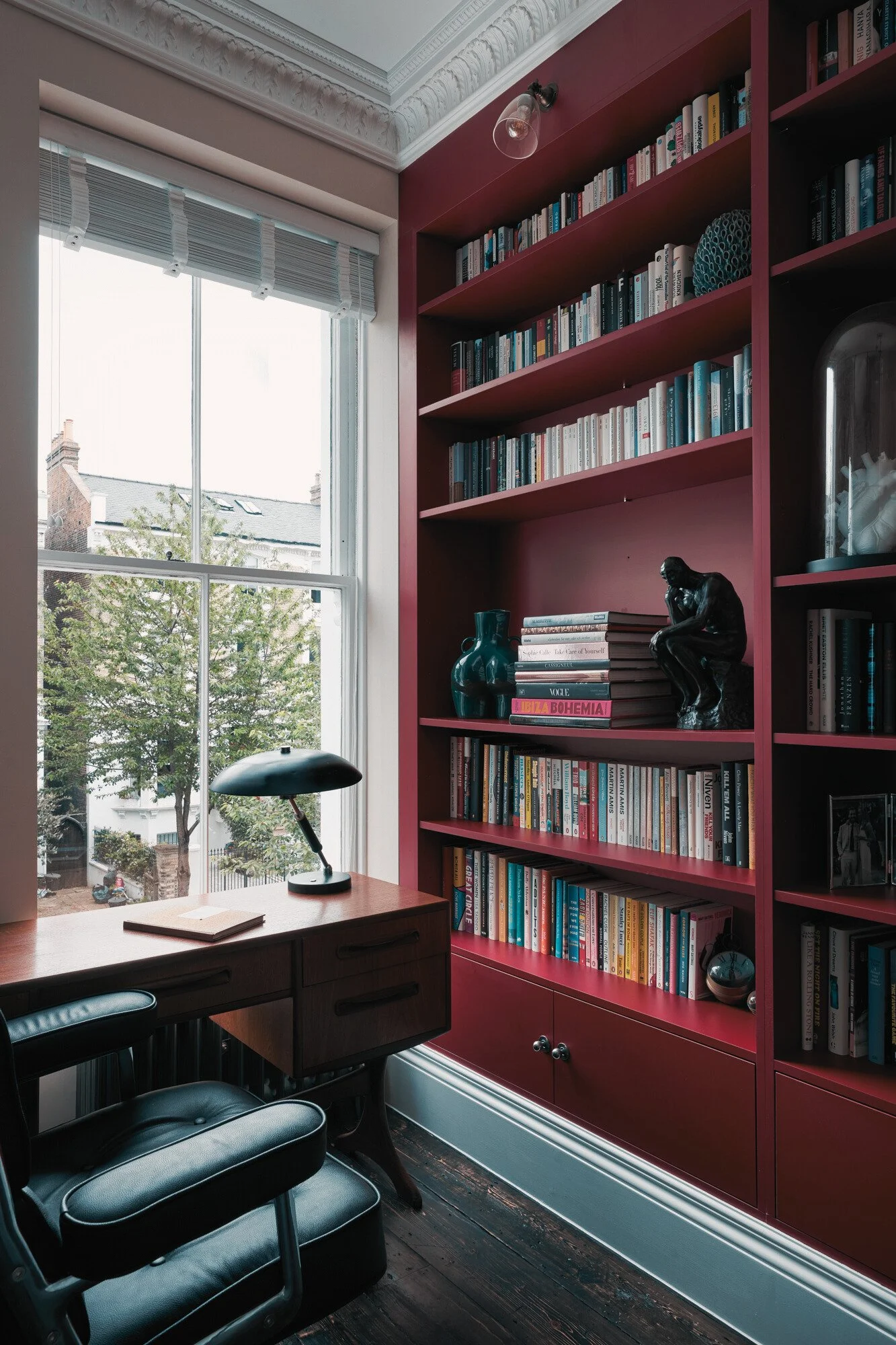Interior of a home office with a large pink bookshelf filled with books, a dark wooden desk, a black leather chair, a window with white blinds, and a decorative lampshade.