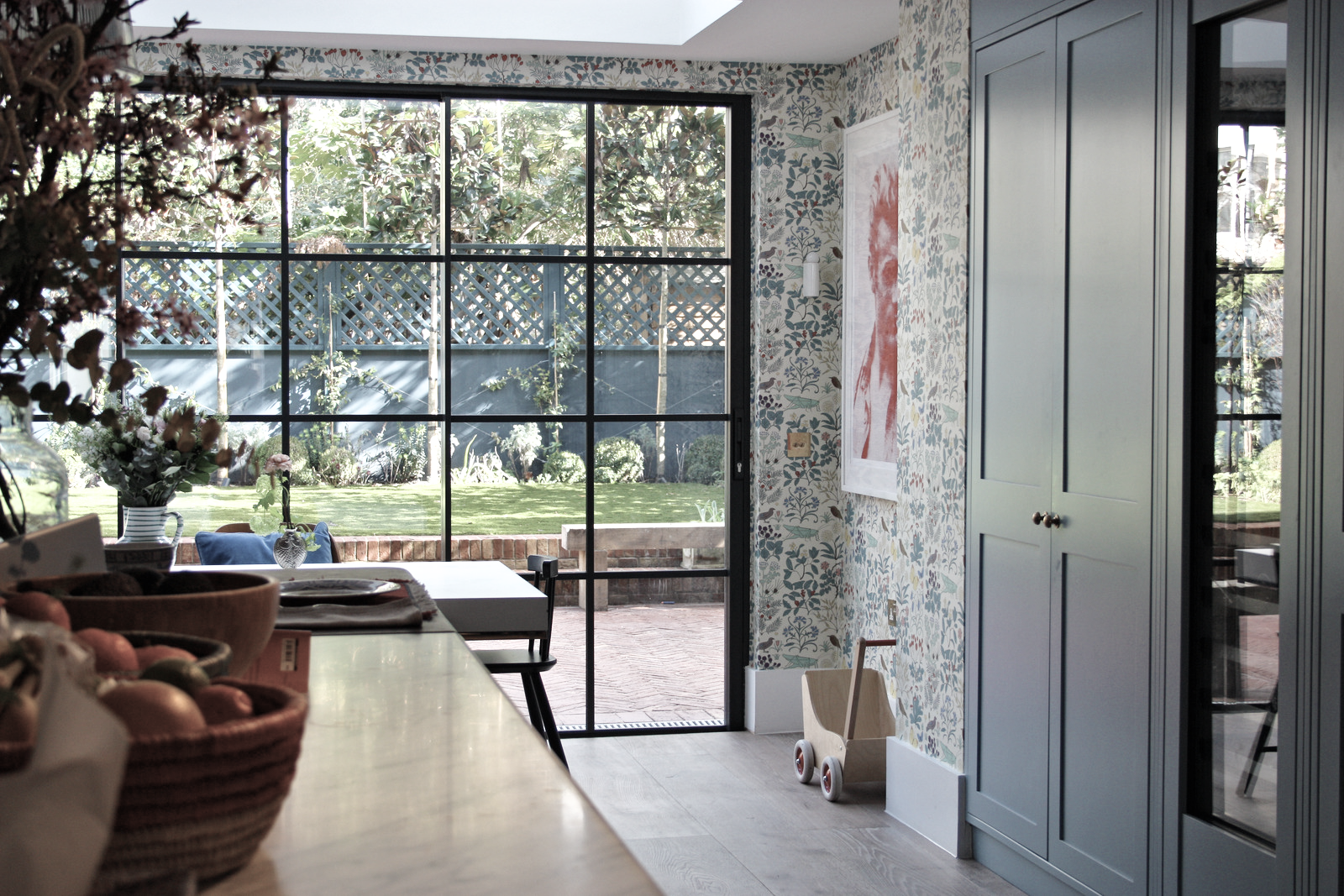 Interior view of a kitchen with a floral wallpaper, a light blue cabinet, and a sliding glass door leading to a backyard with a lawn and trees.