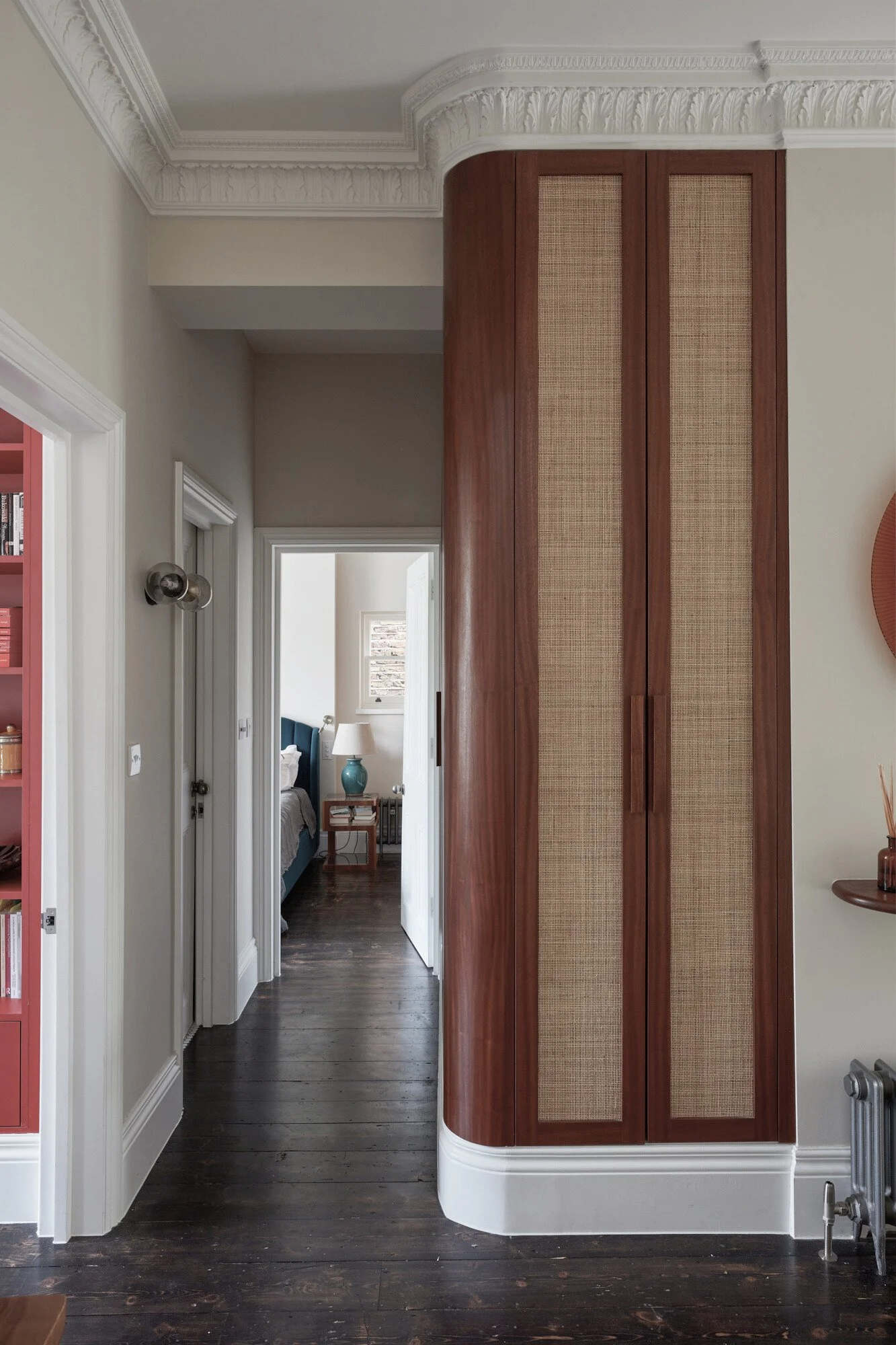 Interior view of a home hallway with dark wooden floors, white walls, ornate crown molding, and a built-in curved wooden cabinet with woven panel doors. A glimpse into a bedroom with a blue headboard, white curtains, and a bedside table with a blue l