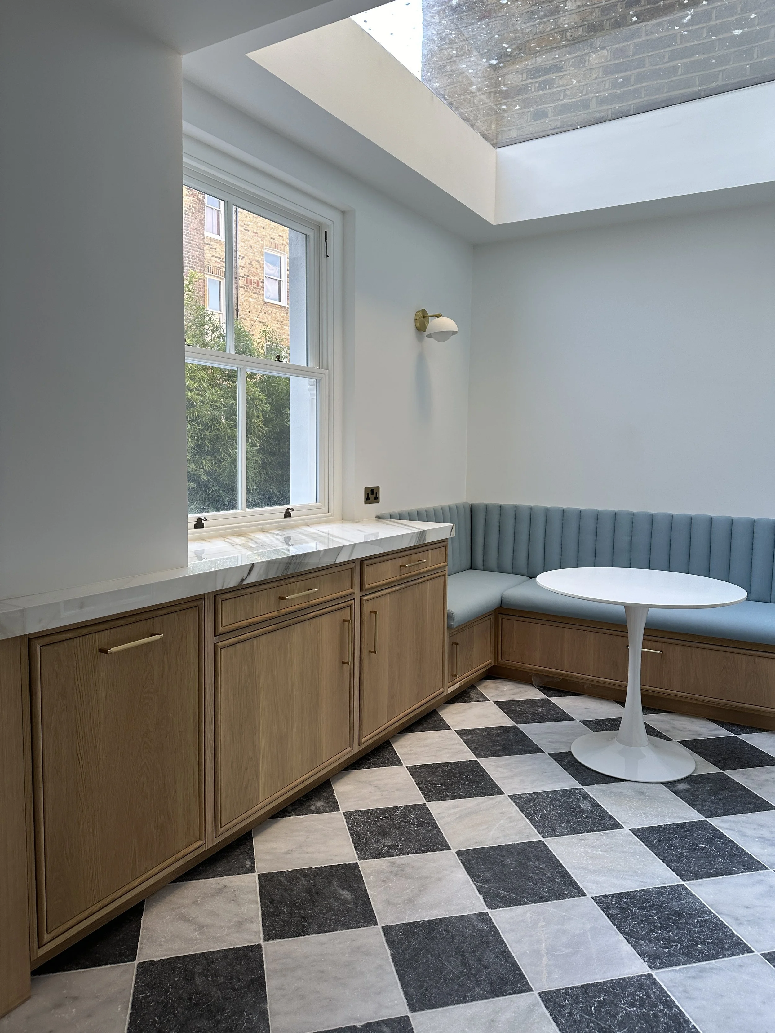 Corner of a room with a large window, wooden cabinets beneath it, and a built-in curved cushioned bench with a white round table in front. The floor has a black and white checkered pattern, and a wall with a sconce light is visible.