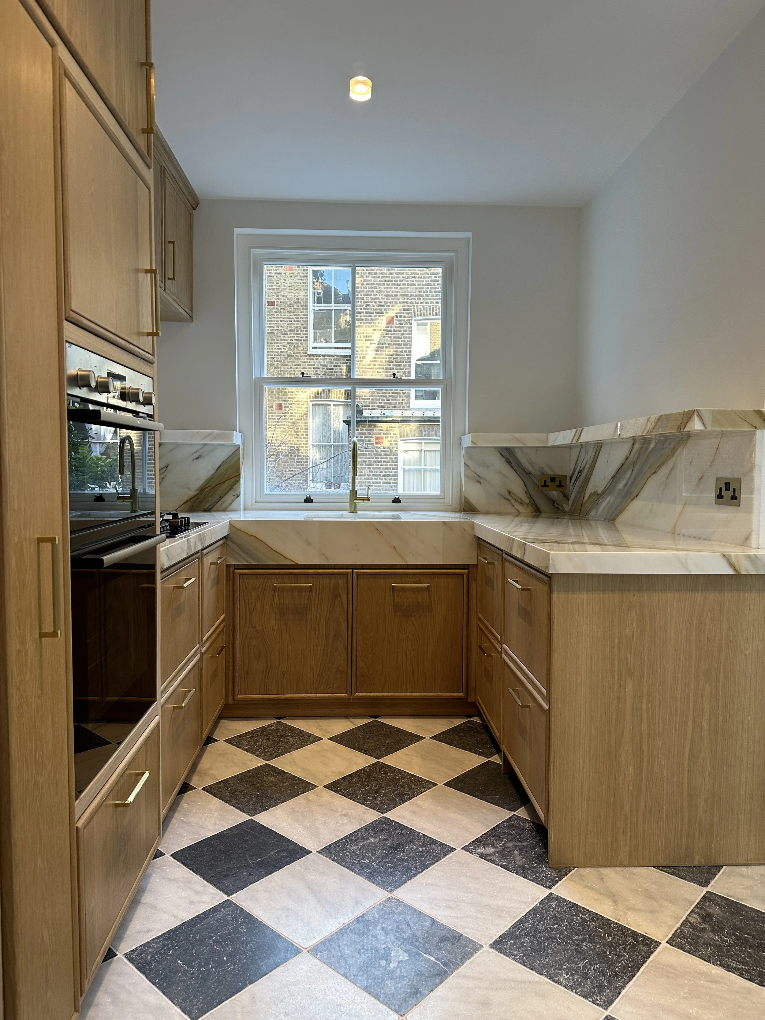 Kitchen with wooden cabinets, marble countertops, and a black and white checkered tile floor. A window over the sink shows brick buildings outside.