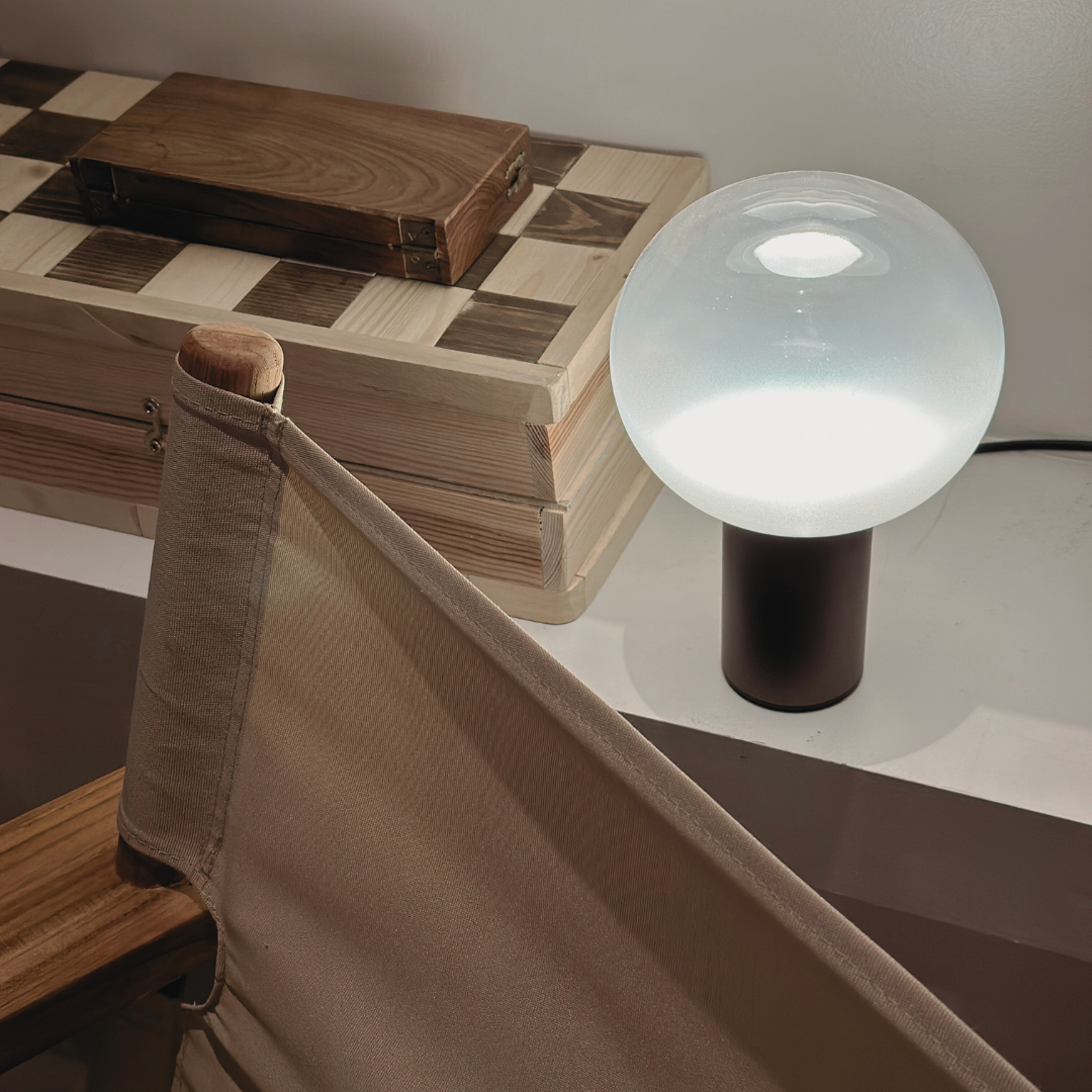 A table with a checkered wooden top, a beige fabric chair, a white lamp with a black base, and a wooden box.