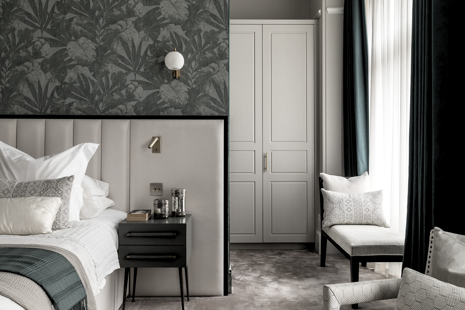 A modern bedroom with a white upholstered headboard, dark nightstand with two candle holders and a book, black wall-mounted reading light, floral dark wallpaper above the headboard, built-in white wardrobe, beige chair by a window with dark curtains,