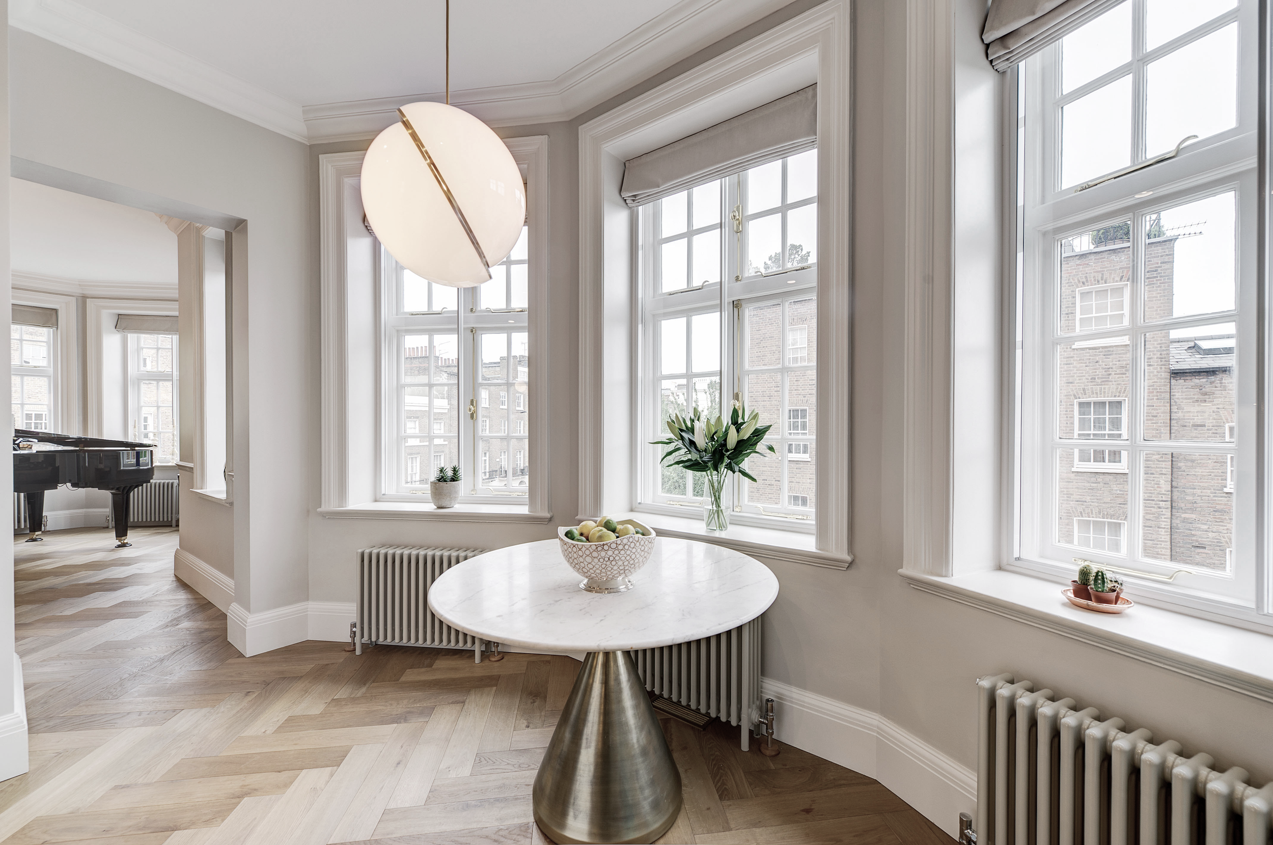 Bright room with large windows, a marble-topped table with a brass base, a bowl of apples, a vase of lilies, and small potted plants.