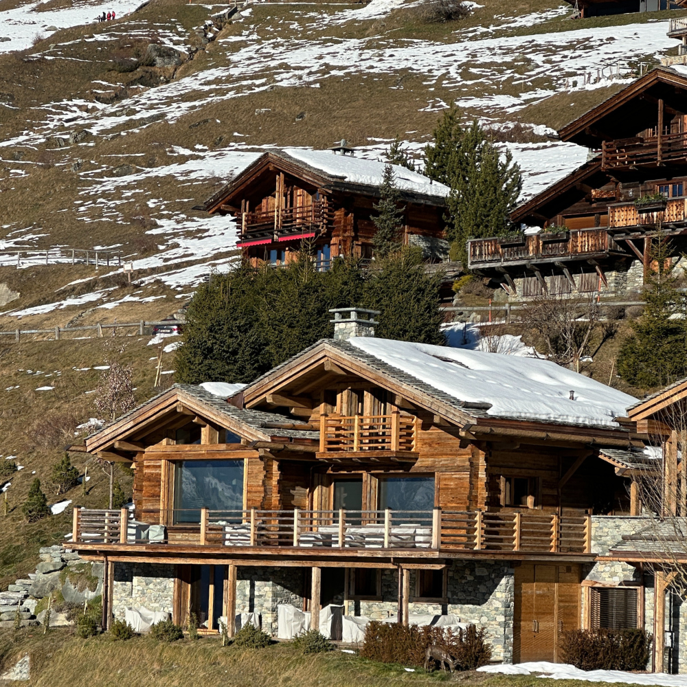 Multiple wooden chalets on a hillside with patches of snow and evergreen trees.