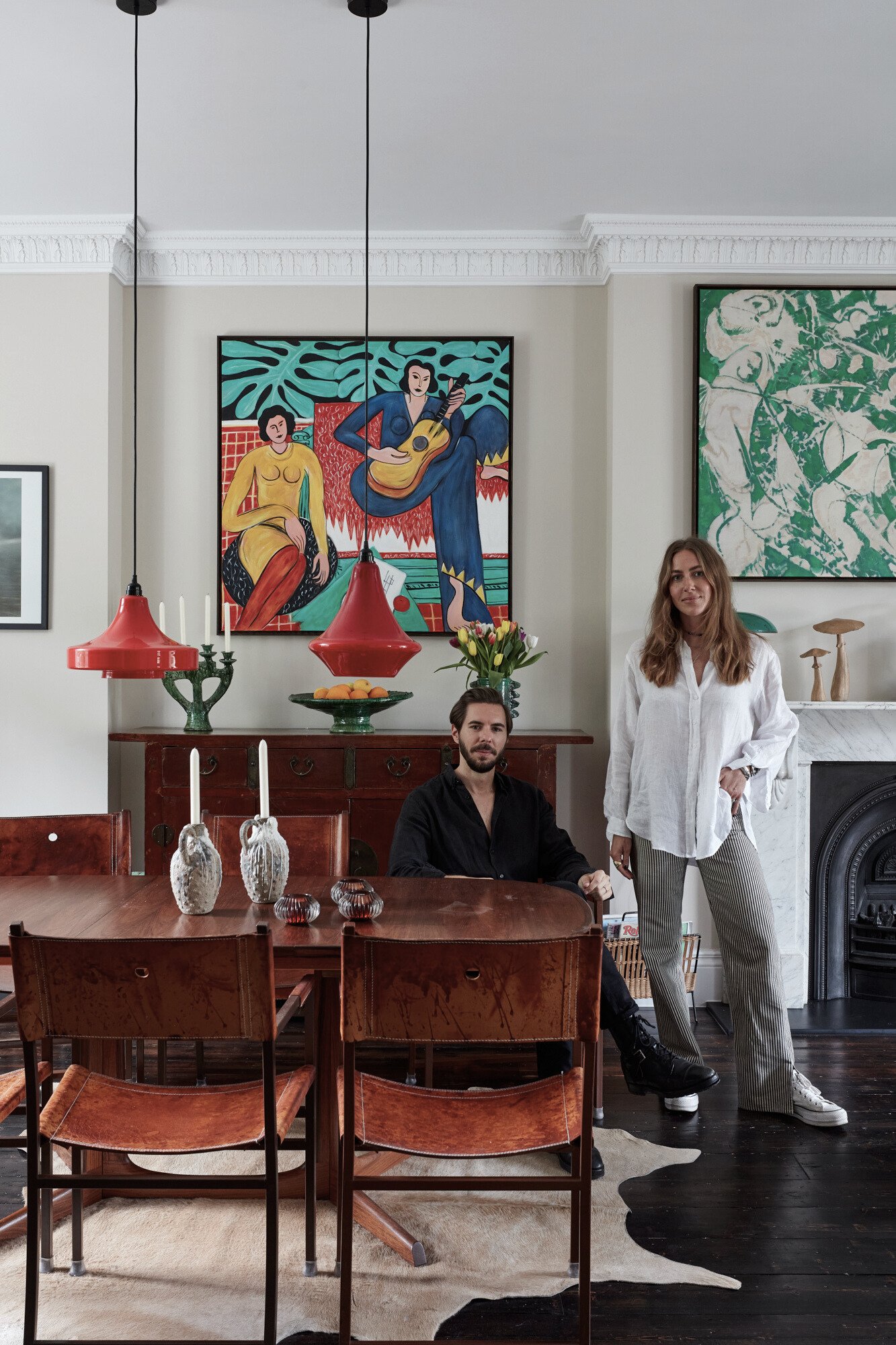 A man and woman in a stylish living room with modern and vintage decor. The man is sitting at a wooden table, and the woman is standing next to him. The room features colorful artwork on the walls, a fireplace, and a dark wood floor with a cowhide ru