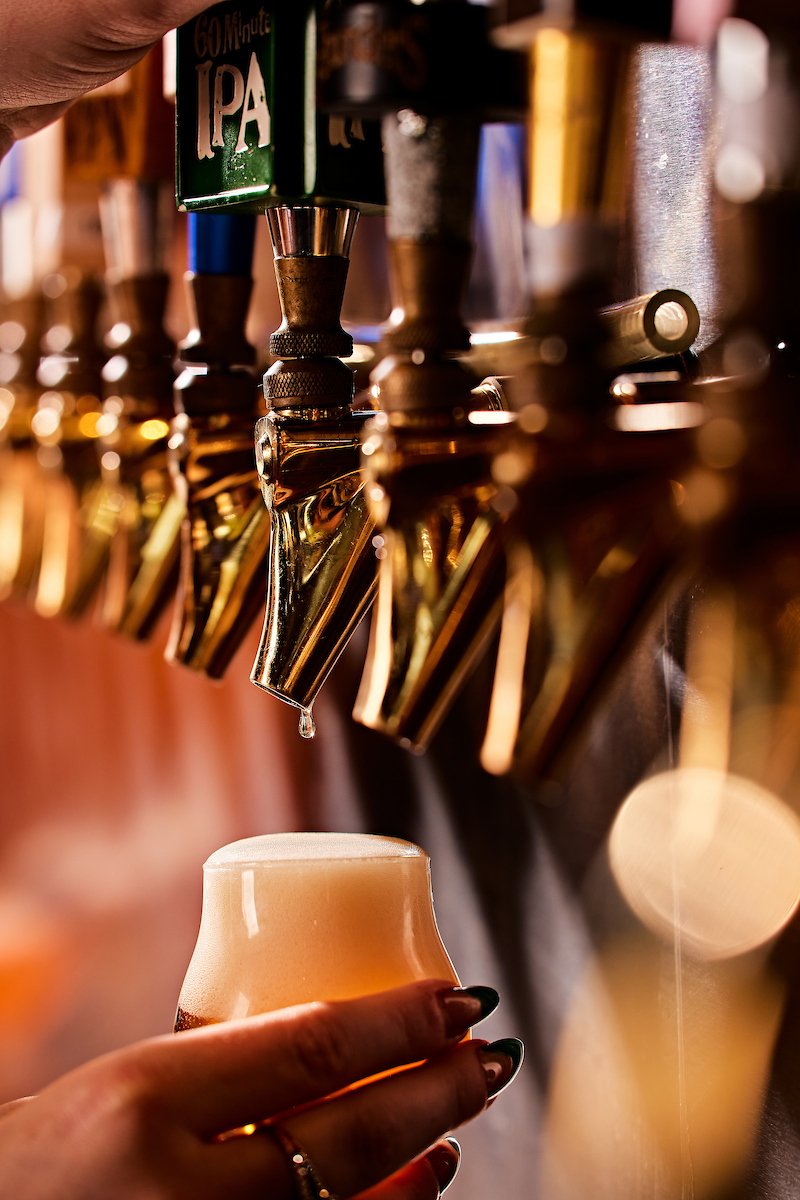 A close-up of a hand filling a glass with beer from a tap at a bar, with several other taps visible in the background.
