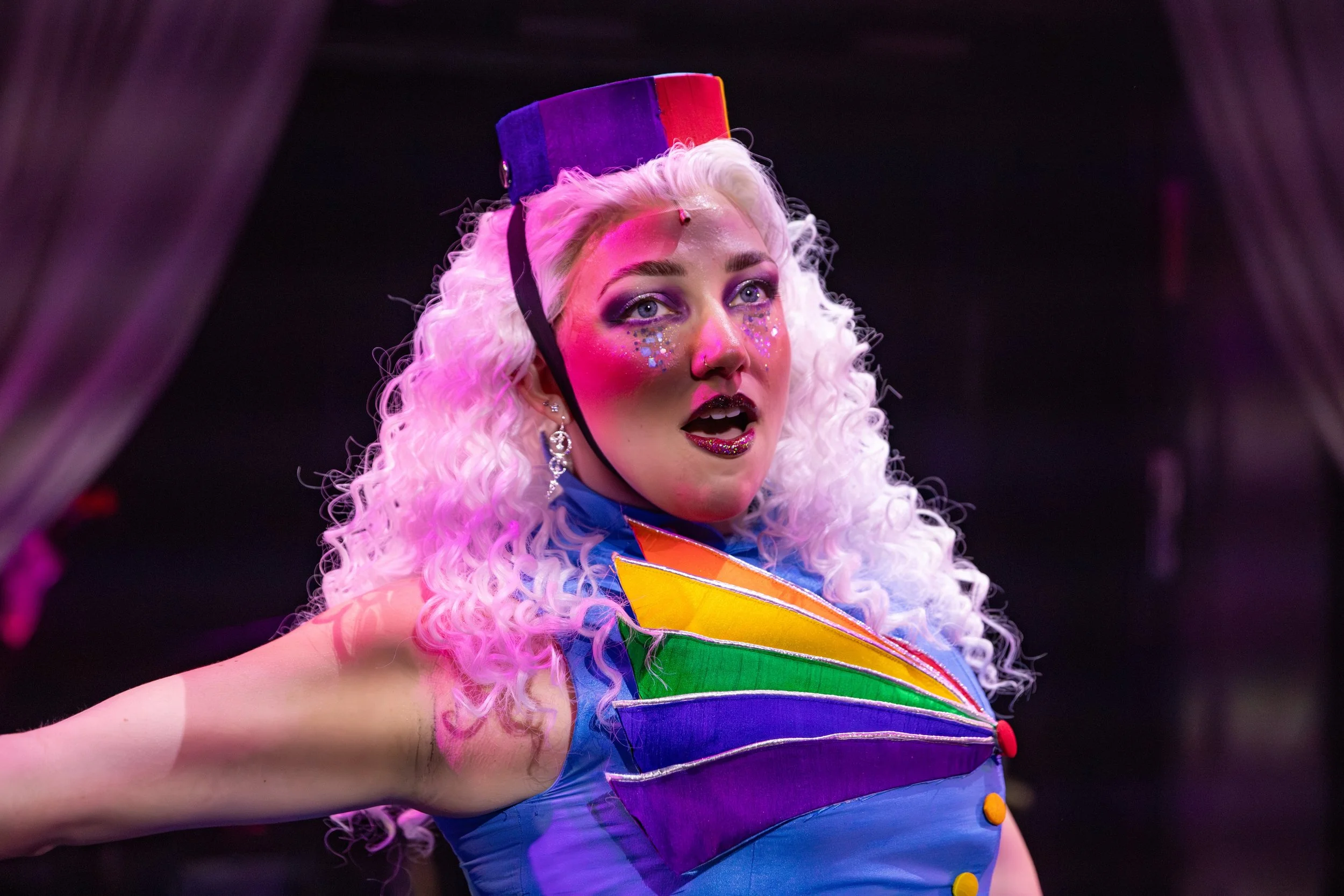 Close-up of a woman with curly white hair wearing a colorful clown costume and hat, with makeup and glitter, in a performance setting.
