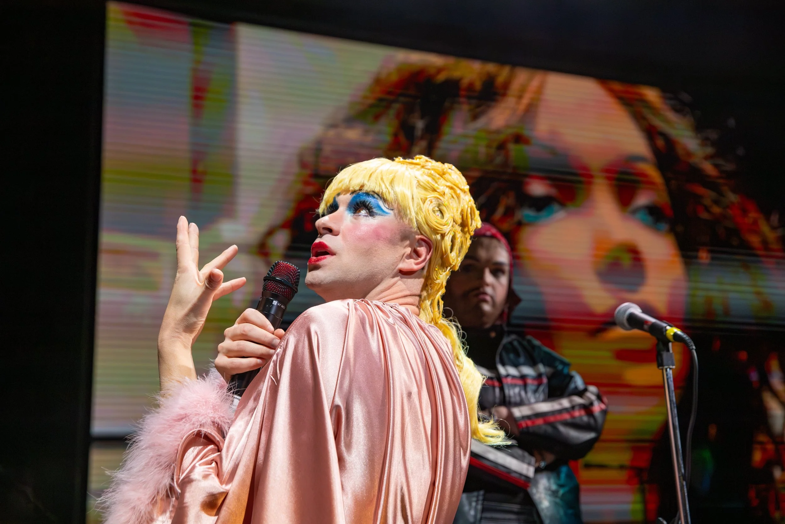 Person with bright yellow hair and colorful makeup holding a microphone, standing in front of a large display screen with a blurred image of a face.