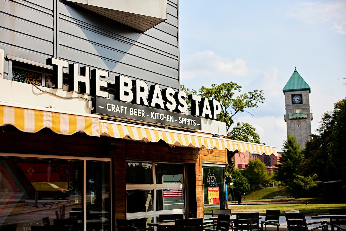 The Brass Tap bar and restaurant with a sign advertising craft beer, kitchen, and spirits, and an outdoor seating area, located in a town with a clock tower visible in the background.
